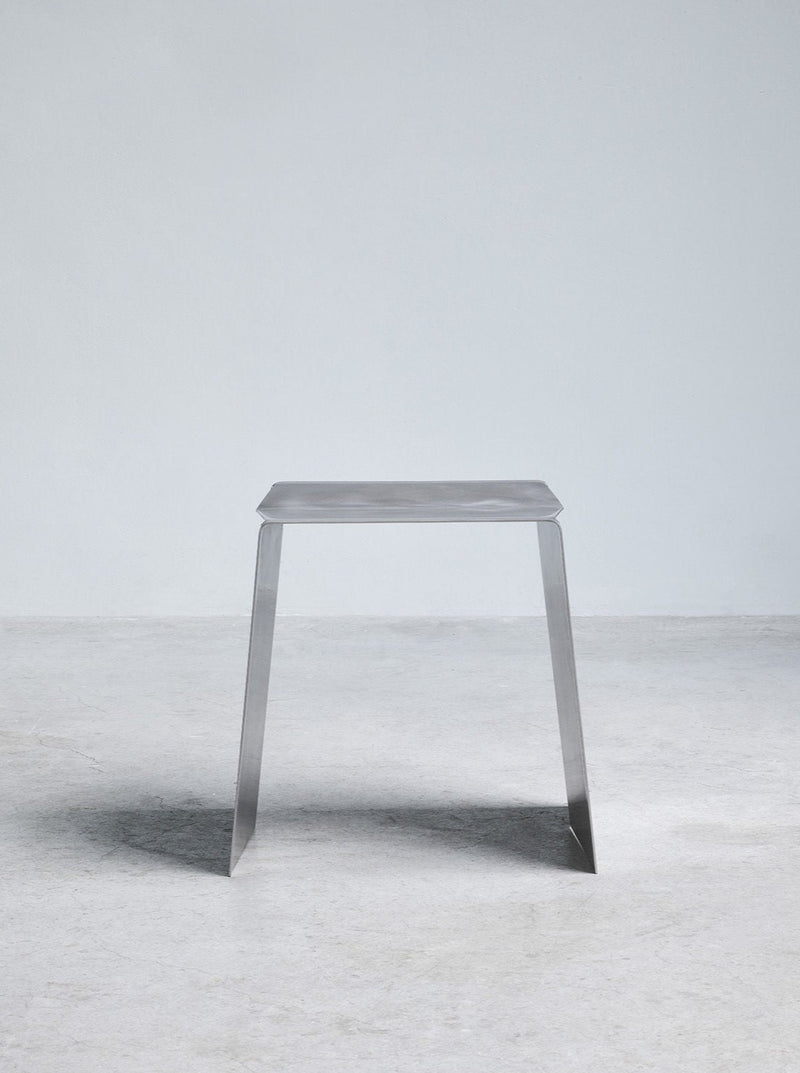 A minimalist Camber Stool by Paul Coenen with angular cuts, standing on a textured gray floor against a simple, light gray background.