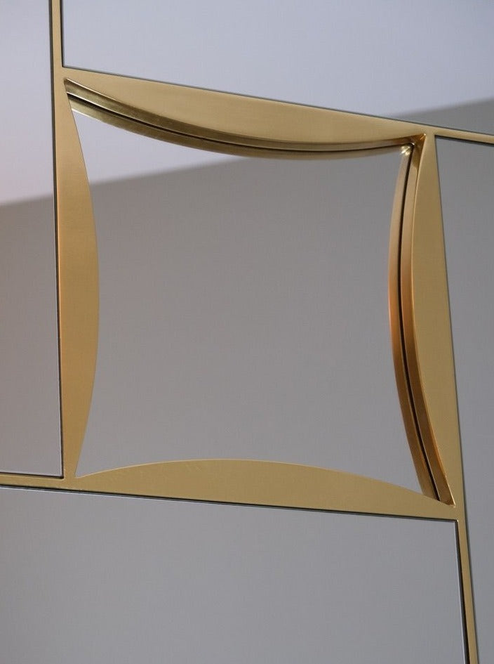 A close-up view of the Mirror Belgo Chrome in Beveled Glass and Brass by LA NOUVELLE GALERIE. The frame consists of geometric shapes with curved and angular lines, creating an abstract and modern design. The decorative element stands out against the blurred, neutral-colored background.