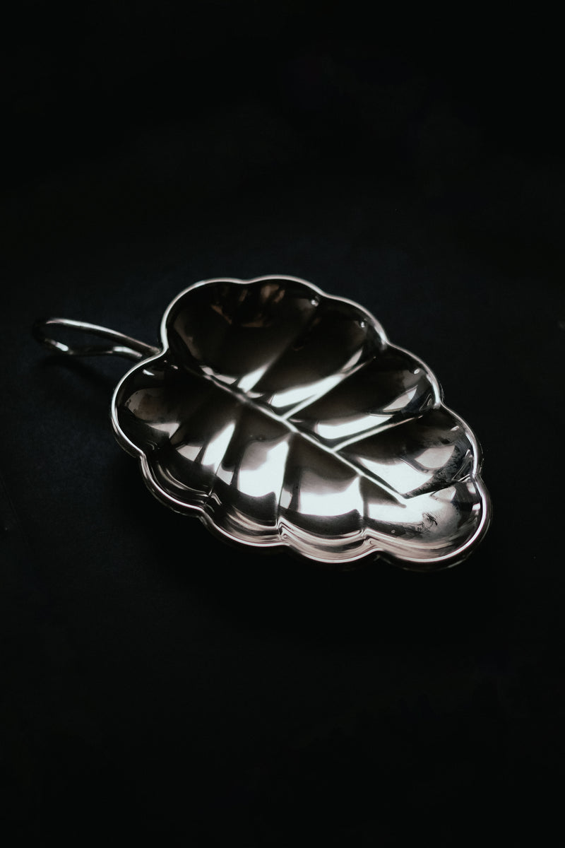 The "Serving Dish Leaf" by septembre studios features a vintage design with intricate detailing, handle, and a shiny silver finish that reflects beautifully on dark surfaces, creating an elegant centerpiece.