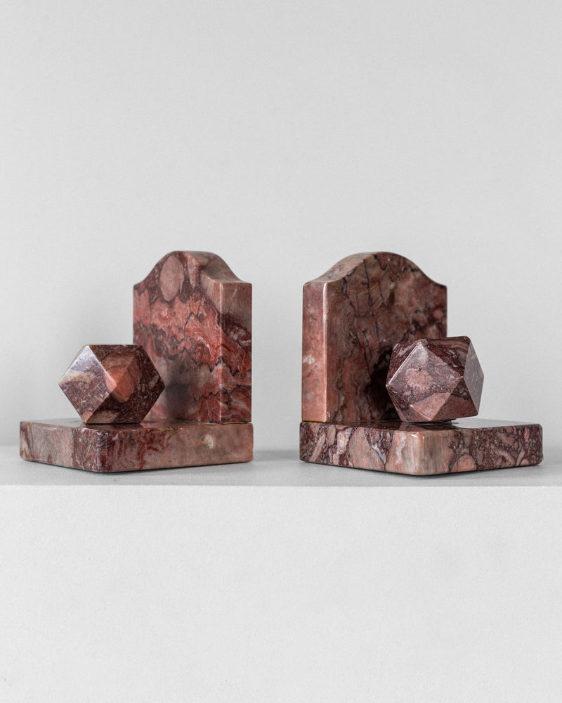 The Spigel Pair of Art Deco Marble Bookends features geometric elements crafted from red marble with intricate patterns, flat bases, and rounded back pieces that create a polyhedral shape. Their elegance is enhanced by a plain light grey background.