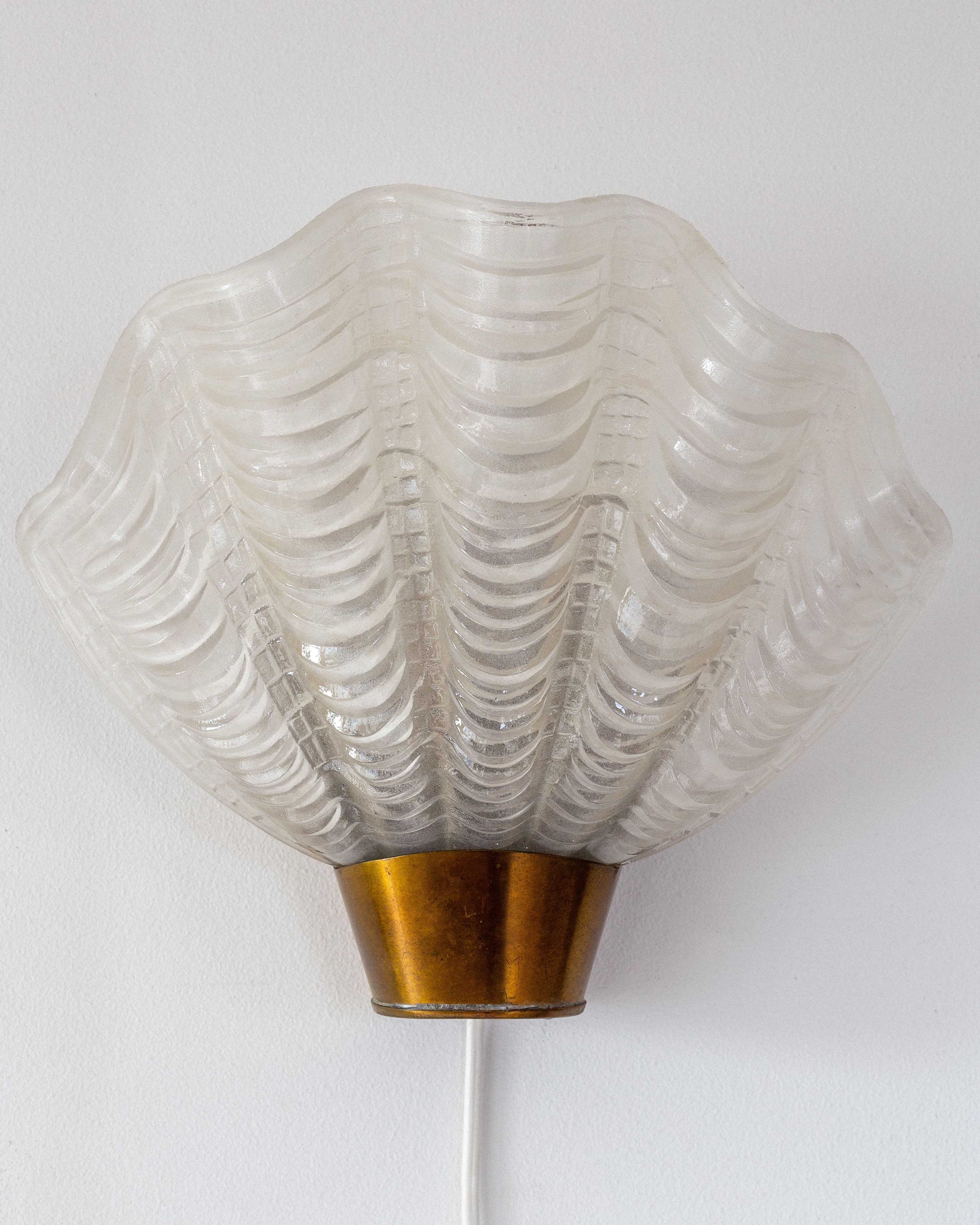 The Spigel Vintage "Coquille" Wall Light from Sweden, 1940s, boasts a seashell-like textured glass shade. Its brass base contrasts with white walls and is complemented by a simple white cord, blending Art Deco vintage charm with modern design.