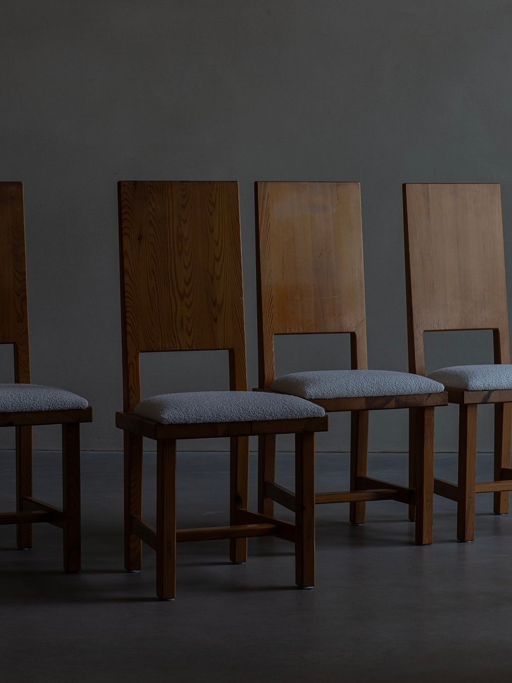 Four Bicci de' Medici Göran Malmvall Model 553 chairs with high backs and light gray cushions are displayed on a dark floor against a dimly lit wall, evoking a moody, minimalist Swedish design atmosphere.