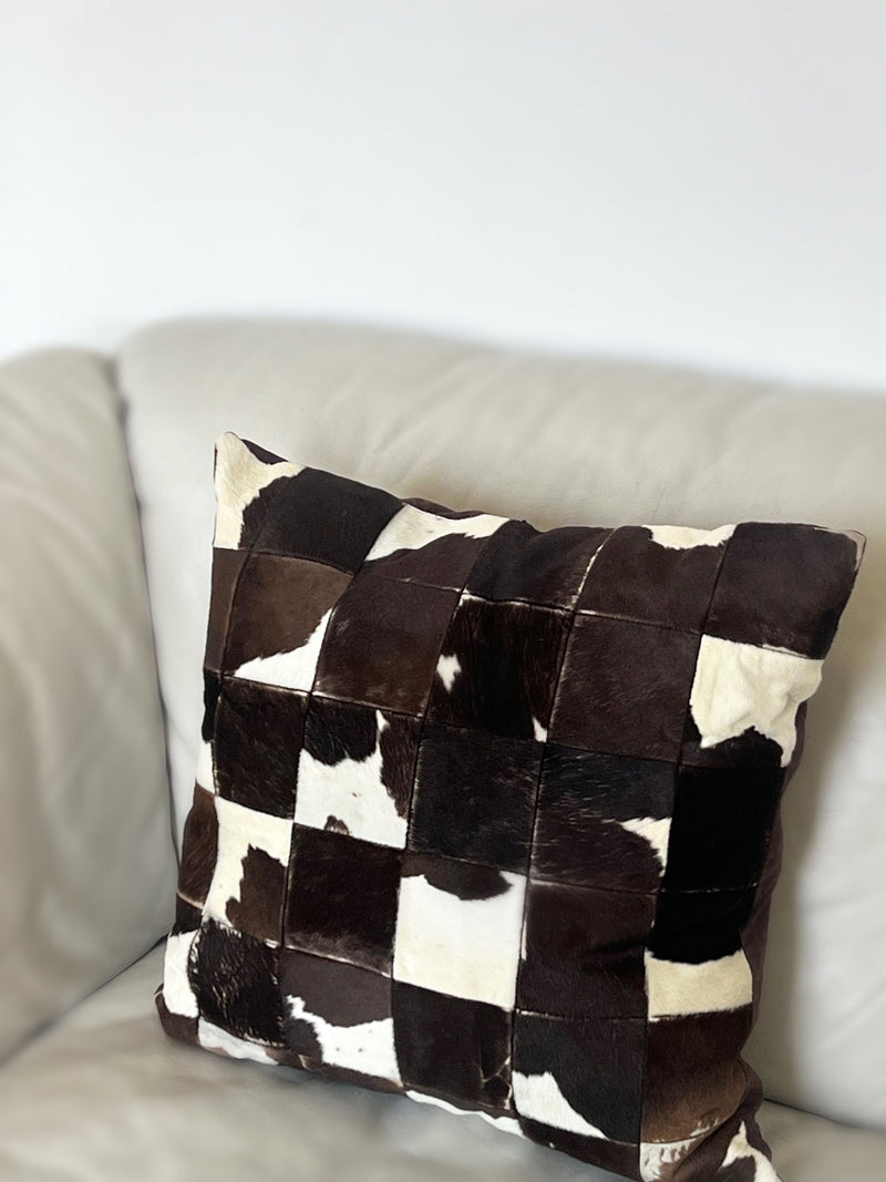 A Porter Studio Cowhide Patchwork Cushion, featuring brown and white cowhide squares with a leather-like texture, adorns a light sofa. Its dark brown linen back contrasts beautifully, while duck feather filling offers supreme comfort.