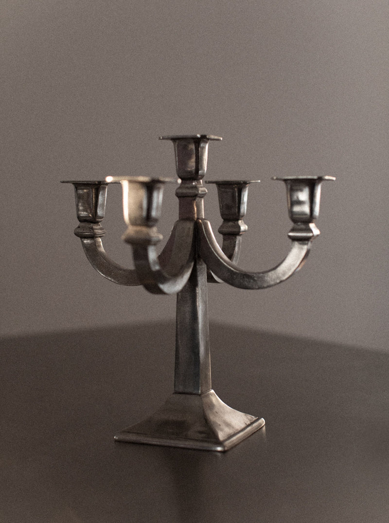 A close-up of the Out For Lunch Mid-century Candleholder with five arms, each featuring an empty candleholder, standing on a dark, smooth surface. The Mid-century Candleholder exudes a distinctive charm with its rustic, slightly tarnished appearance against a neutral gray background, making it a timeless piece of home decor.