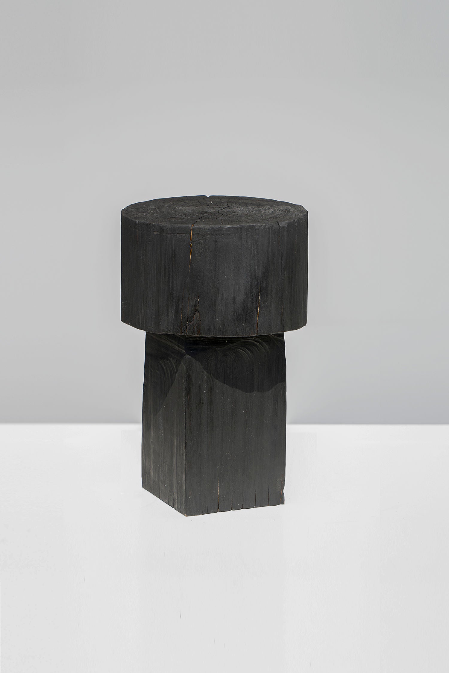 The Burnt Black Coffee Tables/Stools by Touch With Eyes is a minimalist piece, expertly crafted from handmade wood with a round top and square base, shown against a light gray backdrop.