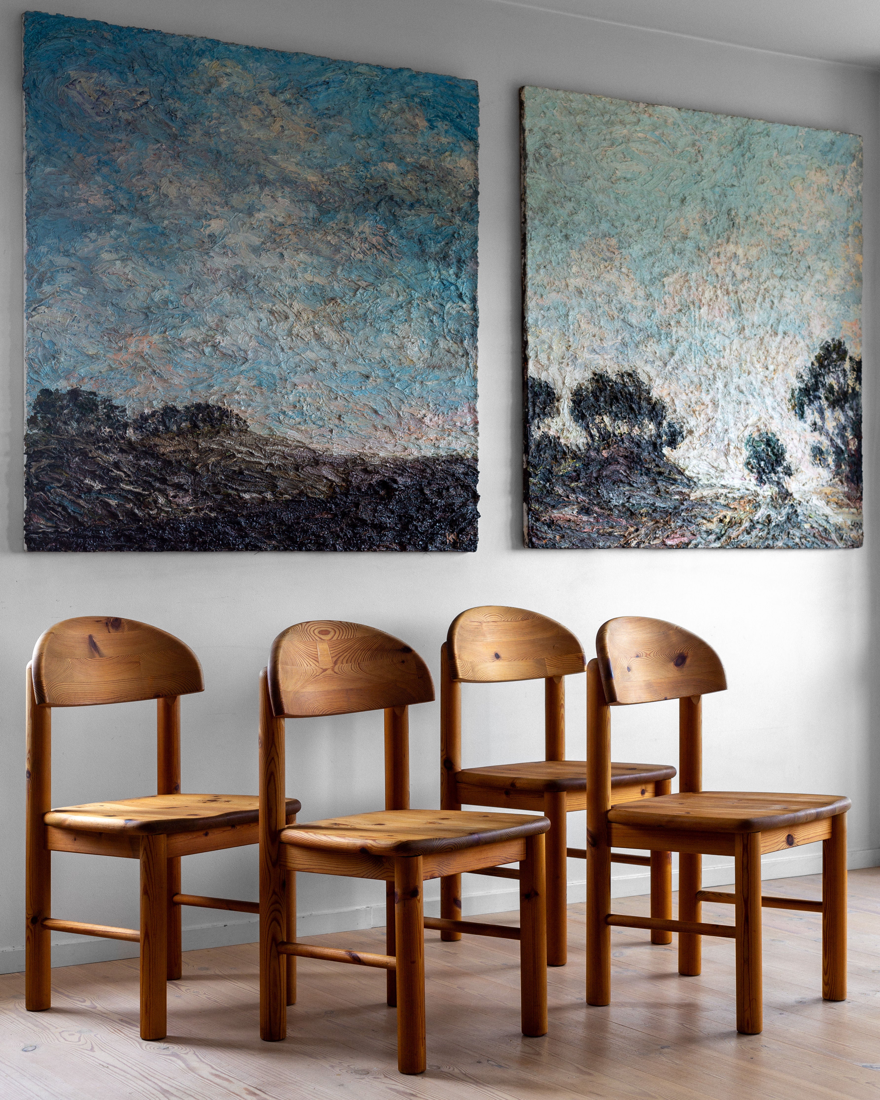 A row of four Spigel's Pine Chairs by Rainer Daumiller, Denmark, 1970s, made of solid pine furniture and showcasing classic Danish design, is placed against a white wall with two large, textured abstract landscape paintings hanging above them. The paintings feature a mix of blue, green, and earthy tones, resembling outdoor scenes with trees and a sky.