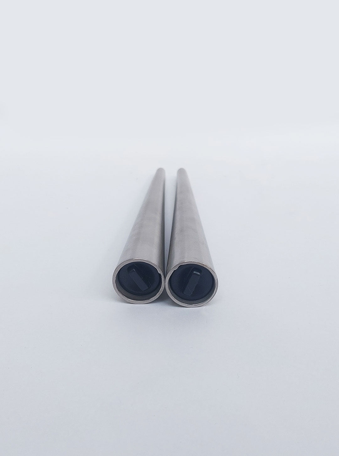 Two cylindrical metal rods are positioned parallel to each other on a white surface. They appear smooth and uniform, with open, darkened ends facing the camera. The background is minimalistic and entirely white, emphasizing the luxurious matte steel finish that exudes elegance and sophistication. Introducing the Conic Pepper and Salt Set by Les Objoies.