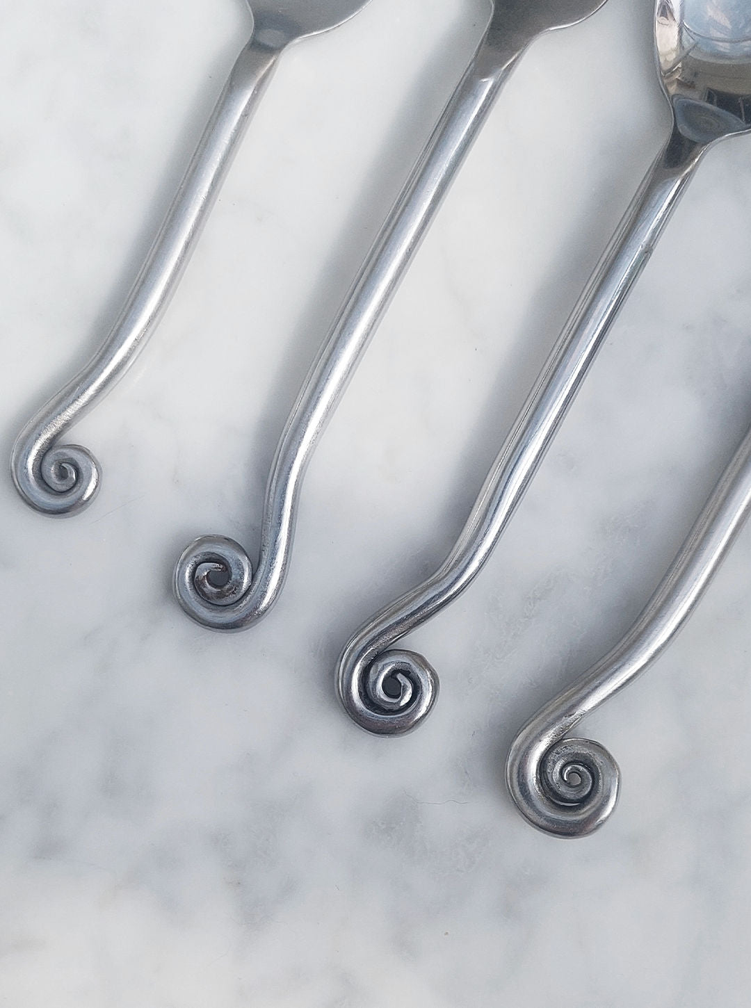 Four forks from the Set of 4 Swirling Cutlery by Les Objoies are arranged on a white marble surface. Each fork features a handle with a decorative spiral design at the end.