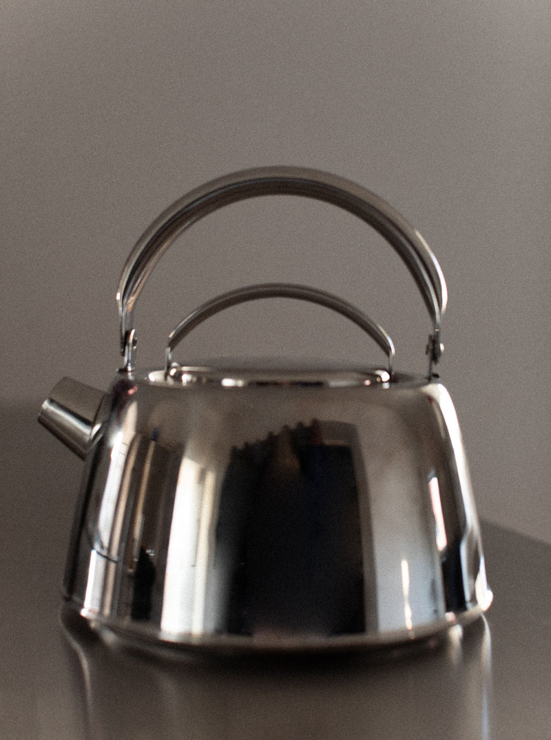 A shiny, chrome **Out For Lunch Chrome Teapot** with a rounded bottom and a sturdy handle reflects light. Placed on a smooth surface with a blurred, neutral-toned background, it enhances the aesthetic appeal of any dining experience by focusing attention on its polished elegance.