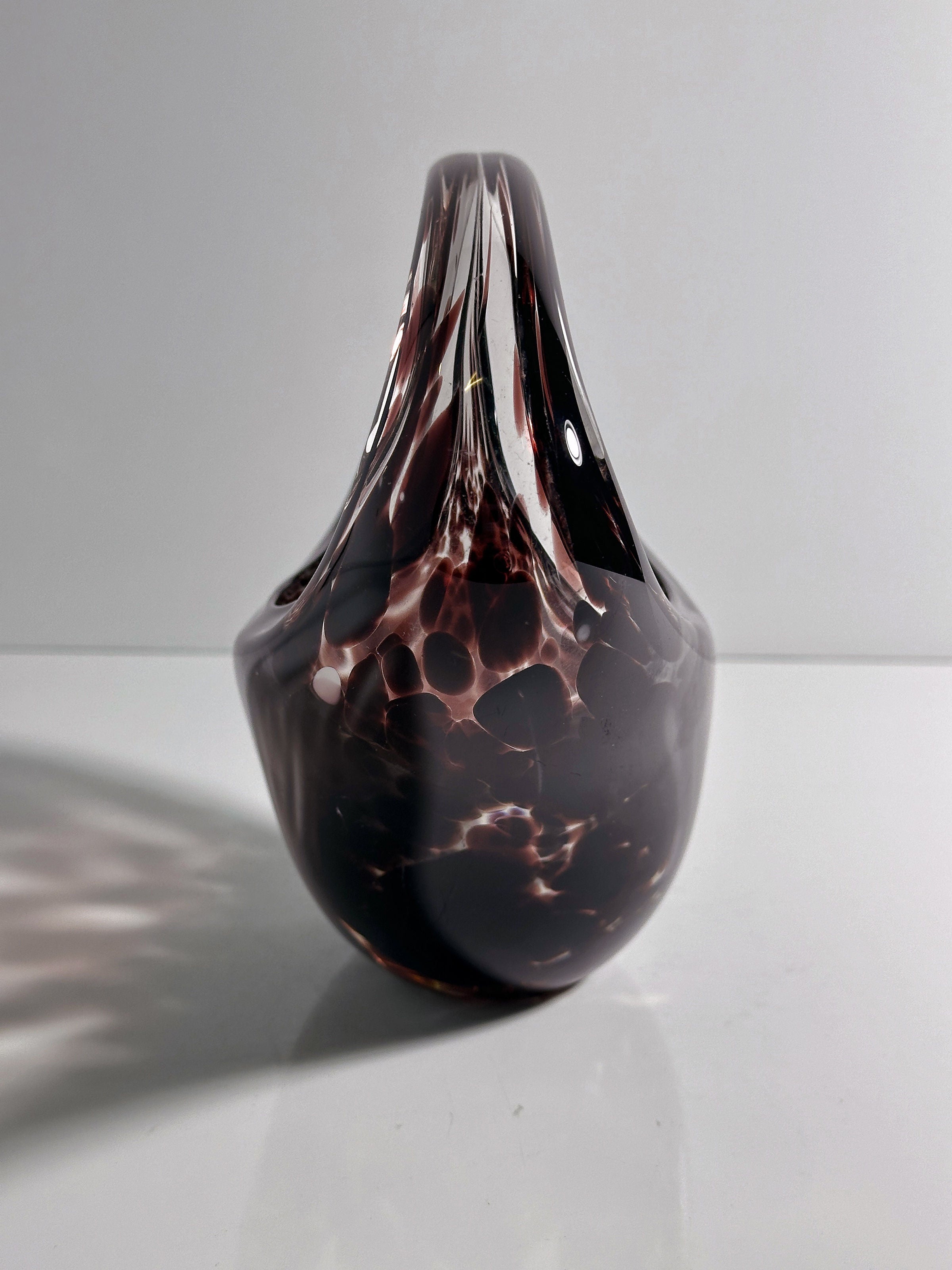 The Murano Tortoise Glass Handle Bowl by Dodo Vintage, with its dark glossy surface and light spots, resembles a stylized vase from the 1970s. Resting on a reflective white surface against a plain gray background, it exudes timeless allure with its flowing edges and abstract elegance.