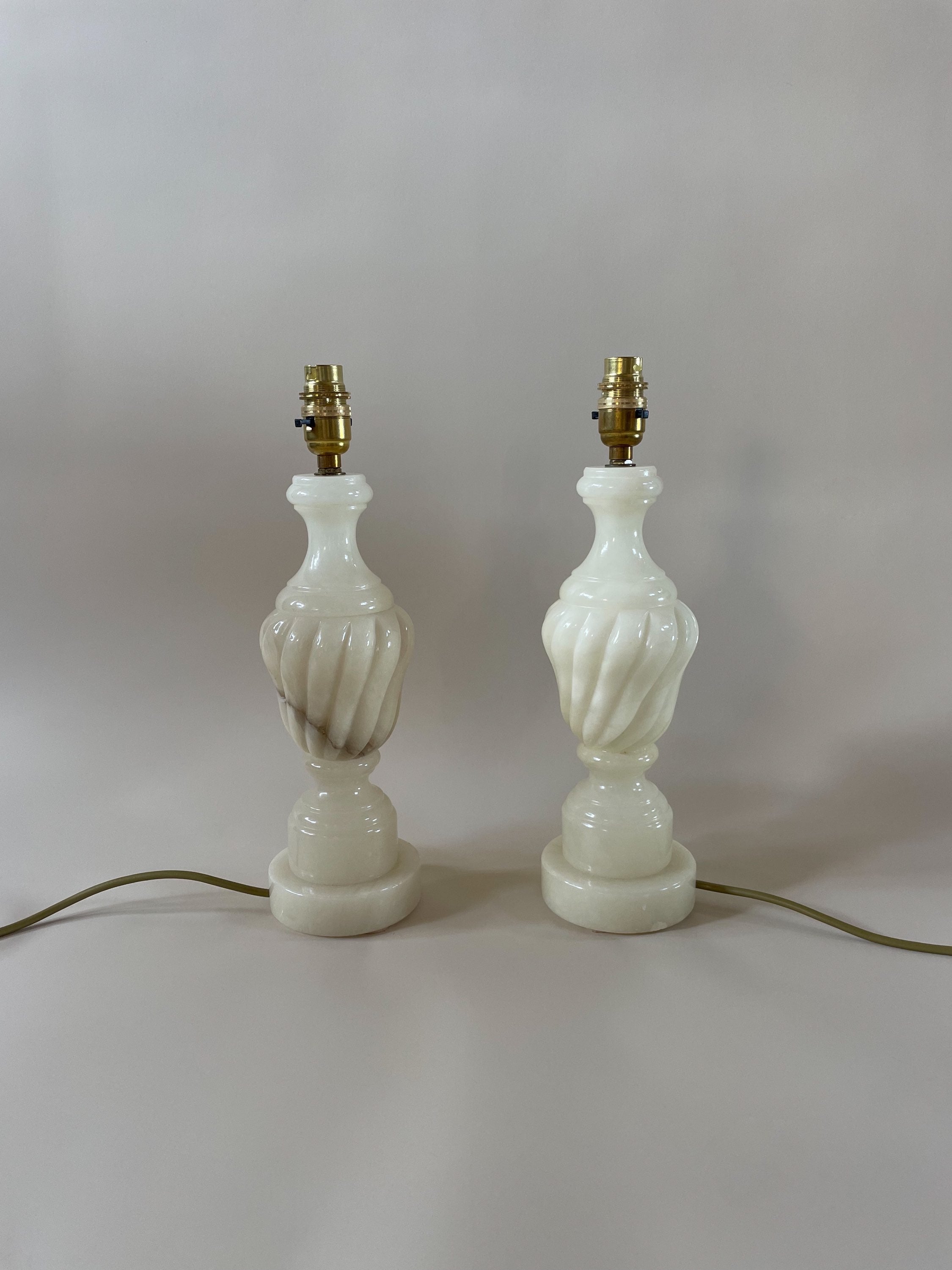 A pair of Maud Vaughan's Pair of Vintage Alabaster Lamps featuring intricately twisted, sculptural white bases. These lamps are shade-free and include brass fittings on top, connected to an antique gold flex cord that rests on a plain, neutral background.