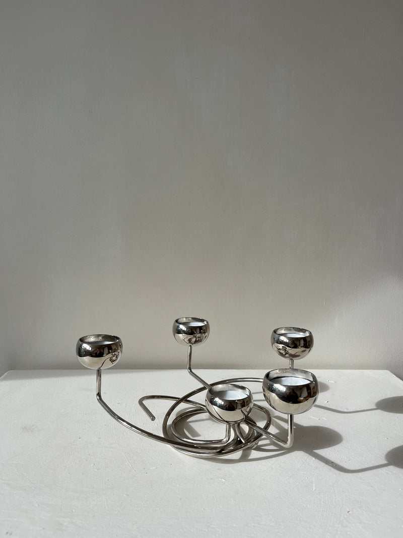 The Chrome Candleholder by septembre studios, featuring a contemporary metallic design with five round holders on a twisted base, sits elegantly on a white surface against a plain light gray background. It casts soft shadows while highlighting its unique angles.