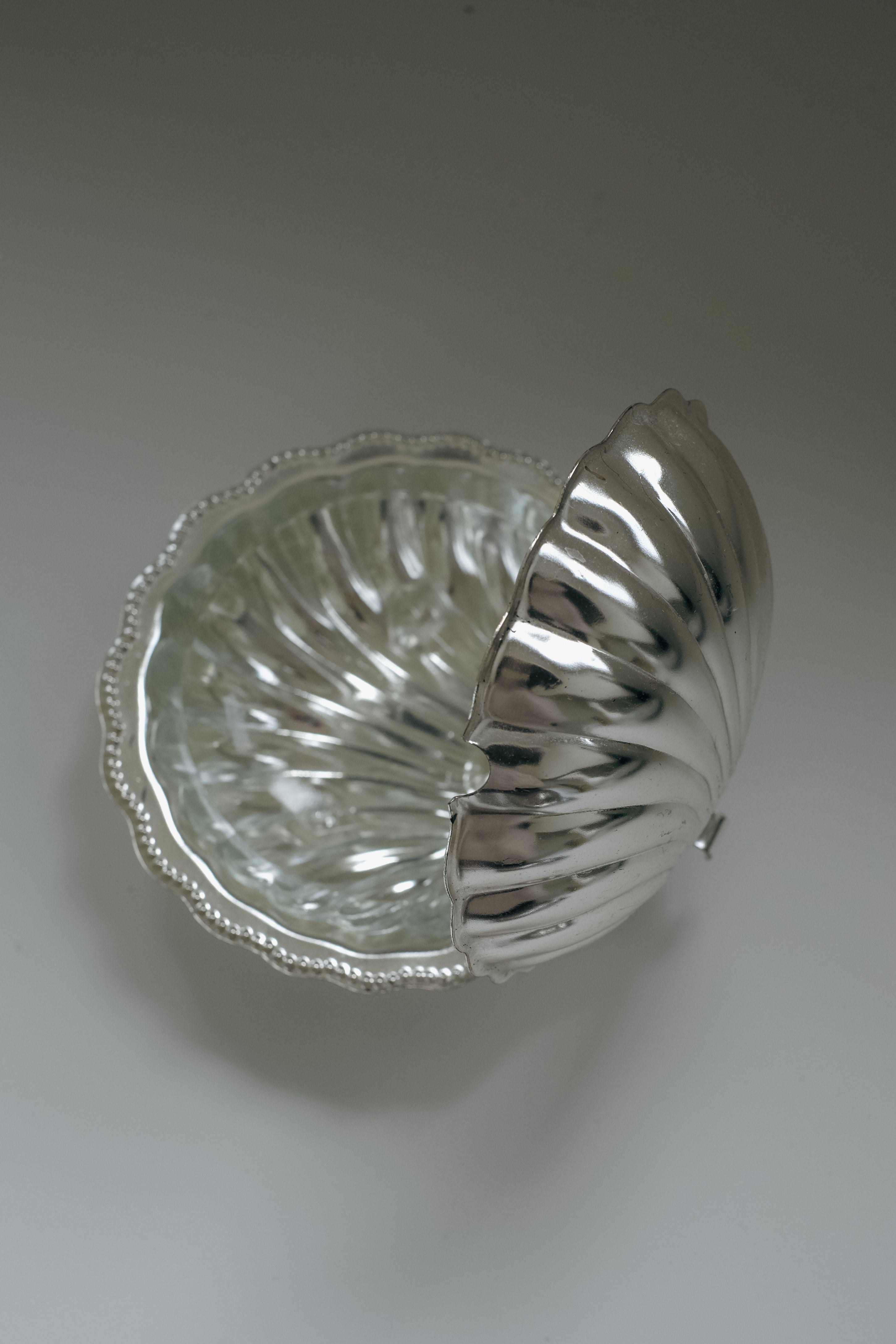 The septembre studios Shell Shaped Butter Dish boasts an elegant ribbed, silver seashell design. A partially open lid reveals a removable glass insert with a matching pattern. Set against a plain white background, its shiny polished surface is striking.