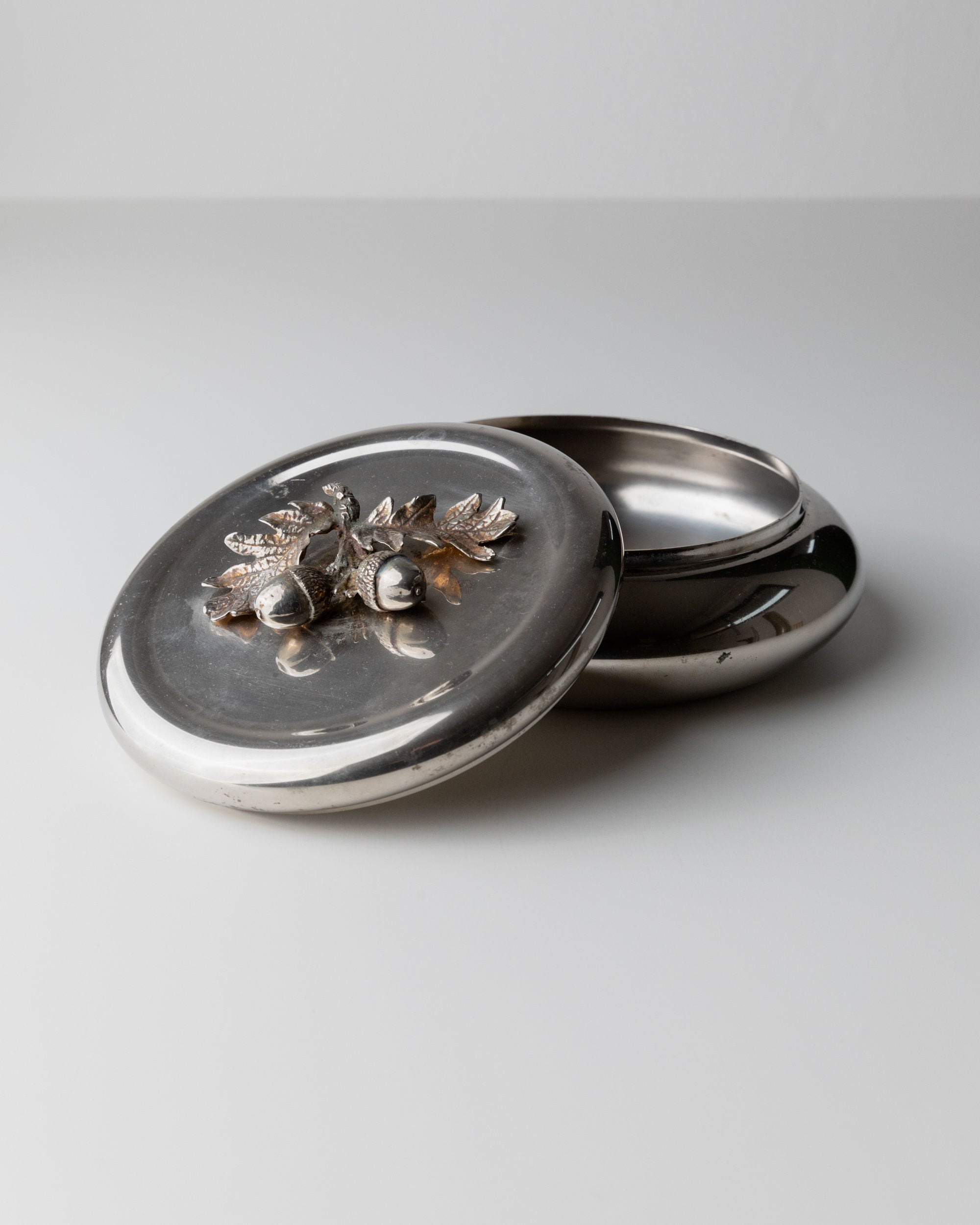 A round, silver-plated Bottega Jacobs box from 1950s Italy features a removable lid adorned with decorative acorns and leaves, complete with an elegant acorn handle. Set on a plain white surface, its slightly ajar lid reveals an empty interior.