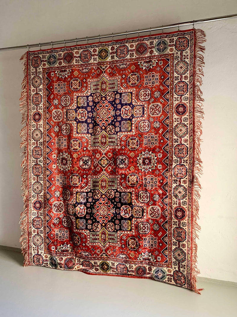 The Veter Vintage "Vintage Silk Oriental Duvet Cover - Rug" features intricate red, blue, and white geometric and floral patterns with fringed edges, displayed vertically against a plain white wall.