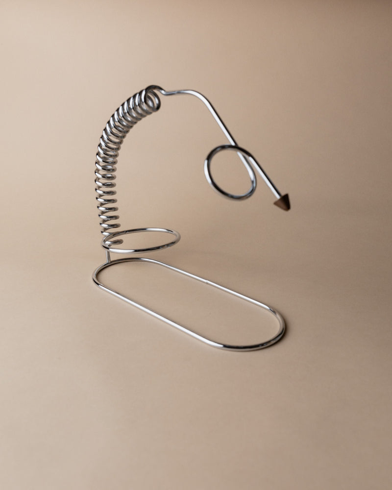 The Bottega Jacobs Bottle Holder, Italy 1980s, features a coiled spring on an oval base with an arrow-tipped loop, offering a touch of space age style against a plain beige background.