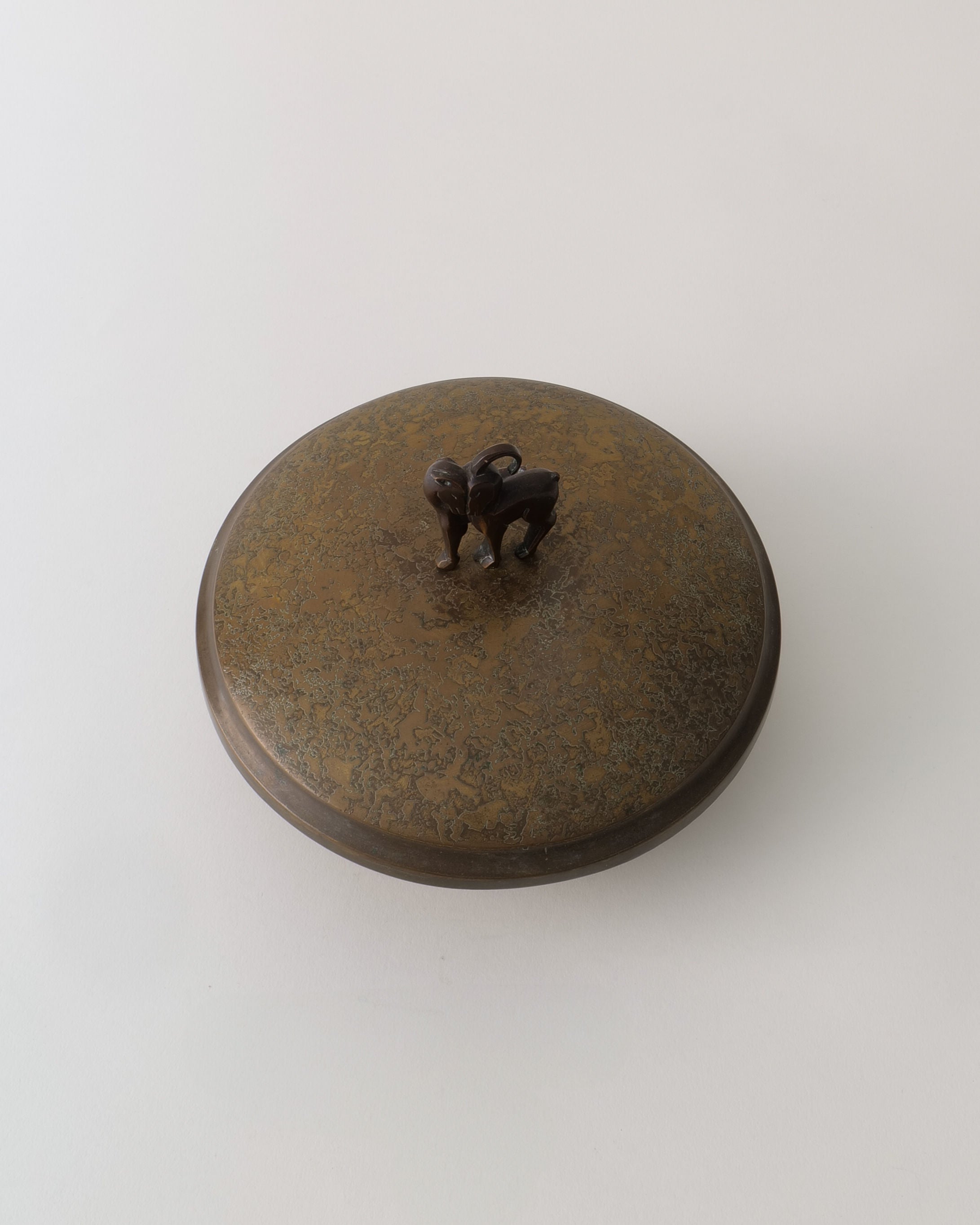 The Deco Box by Pierre Amédée Plasait for Collection apart is a round, bronze-toned Art Deco brass box lid with a textured surface and a sculpted animal handle, reflecting elegant early deco design against a plain light background.