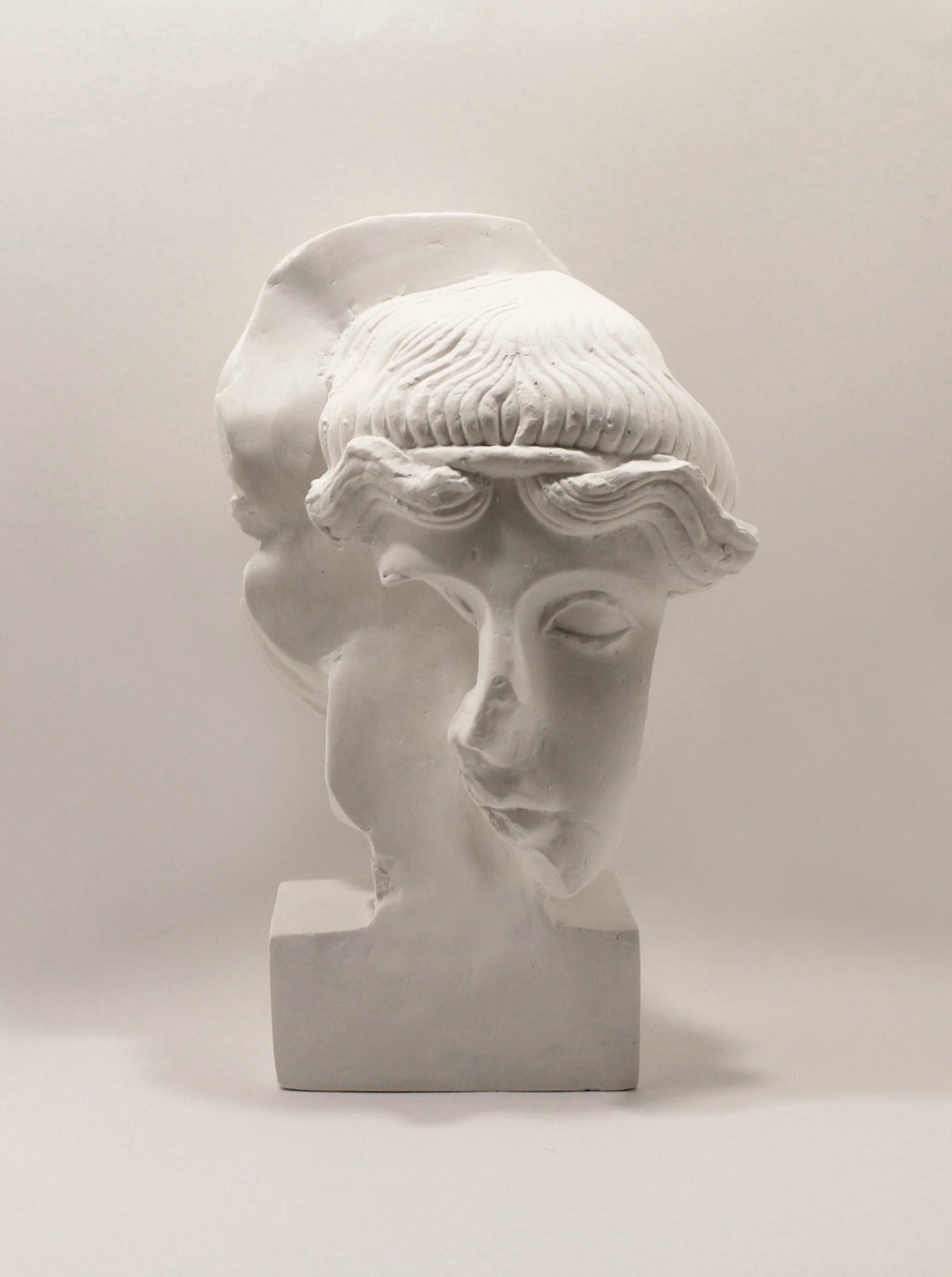 The Fragment #9 Sculpture 2021 by Marina Mankarios is a white plaster bust of a classical female figure, showcasing wavy hair and intricate facial features. This art piece features a clever merge of side and front profiles, set against a plain, light-colored background, exuding timeless elegance.