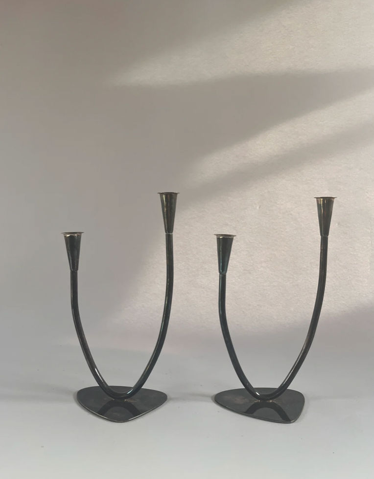 A pair of mid-century candleholders by Maud Vaughan, crafted in Germany, showcases contemporary design with timeless elegance. These sleek black metal pieces feature curved stems and triangular bases, elegantly placed on a light surface against a softly lit background.