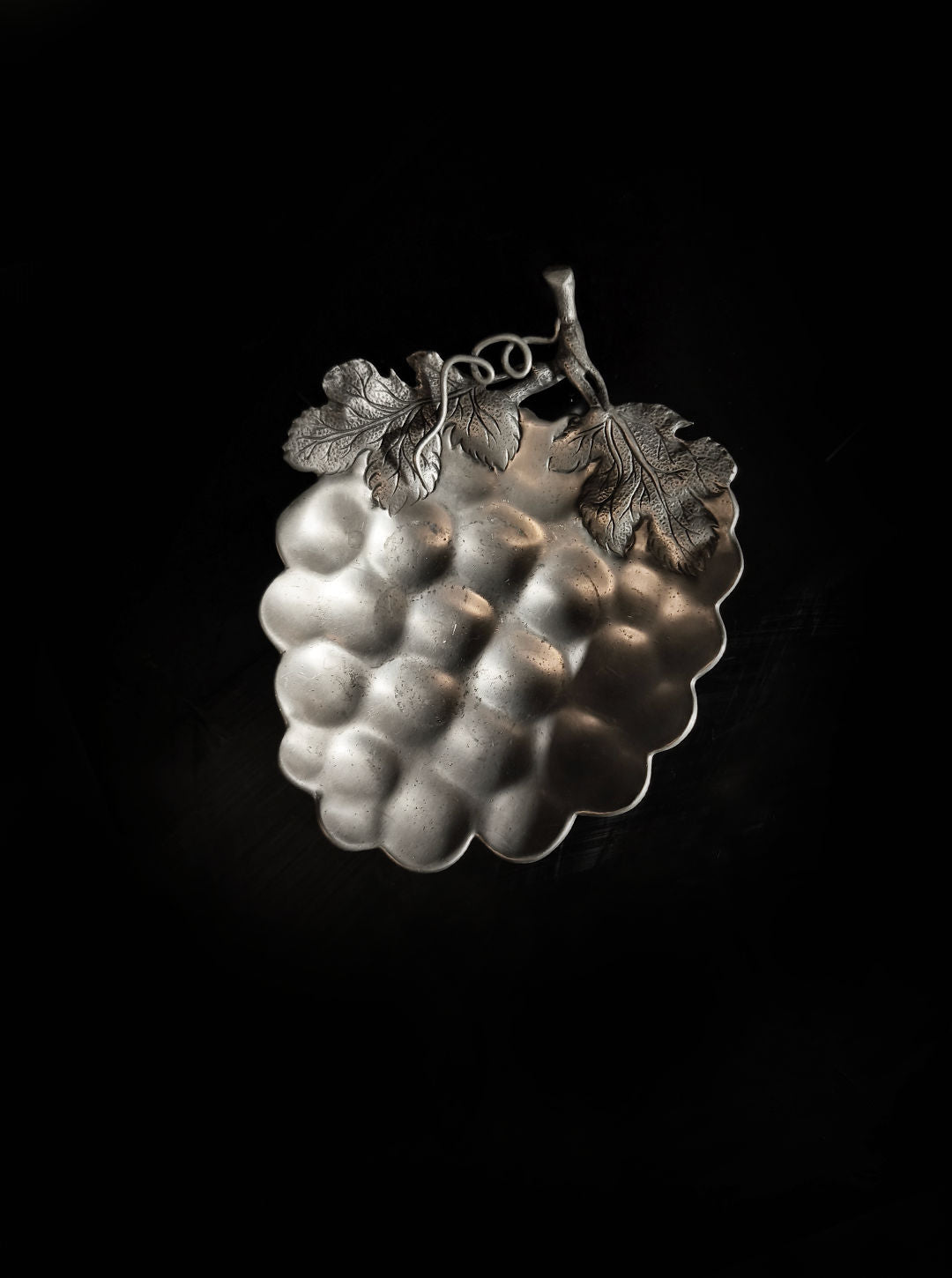 Against a black background, the Grape Vide Poches from Les Objoies displays a silver, grape-shaped dish adorned with intricate leaf designs, highlighting its detailed metalwork and textured surface in a classic grapevine motif. This pewter decorative piece exudes elegance and sophistication in any setting.