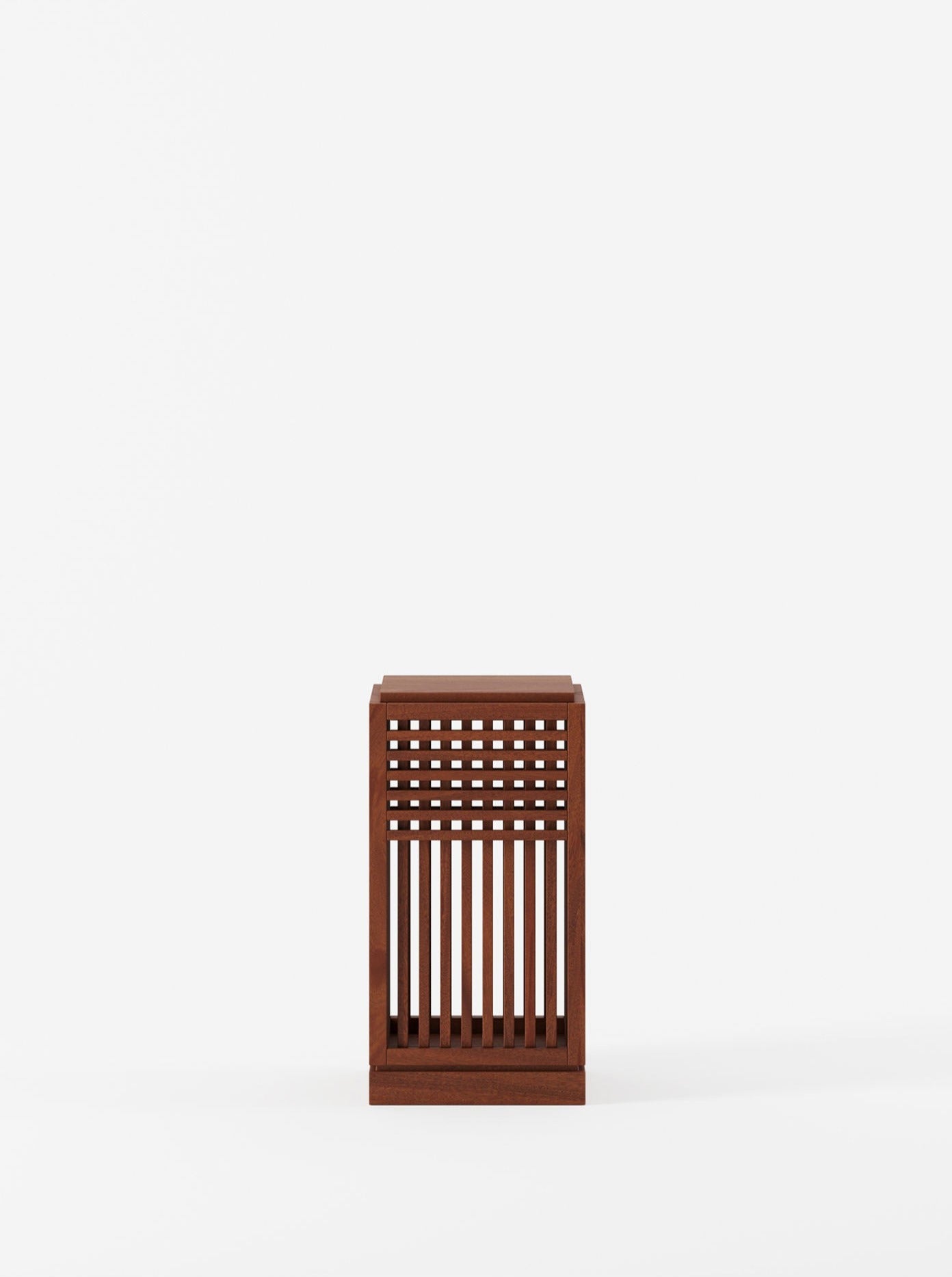 The Riviera Plinth short in Oiled African Mahogany by Lemon is a small, rectangular side table with a lattice design featuring evenly spaced vertical and square cutouts on its sides and front. The table is set against a plain white background.