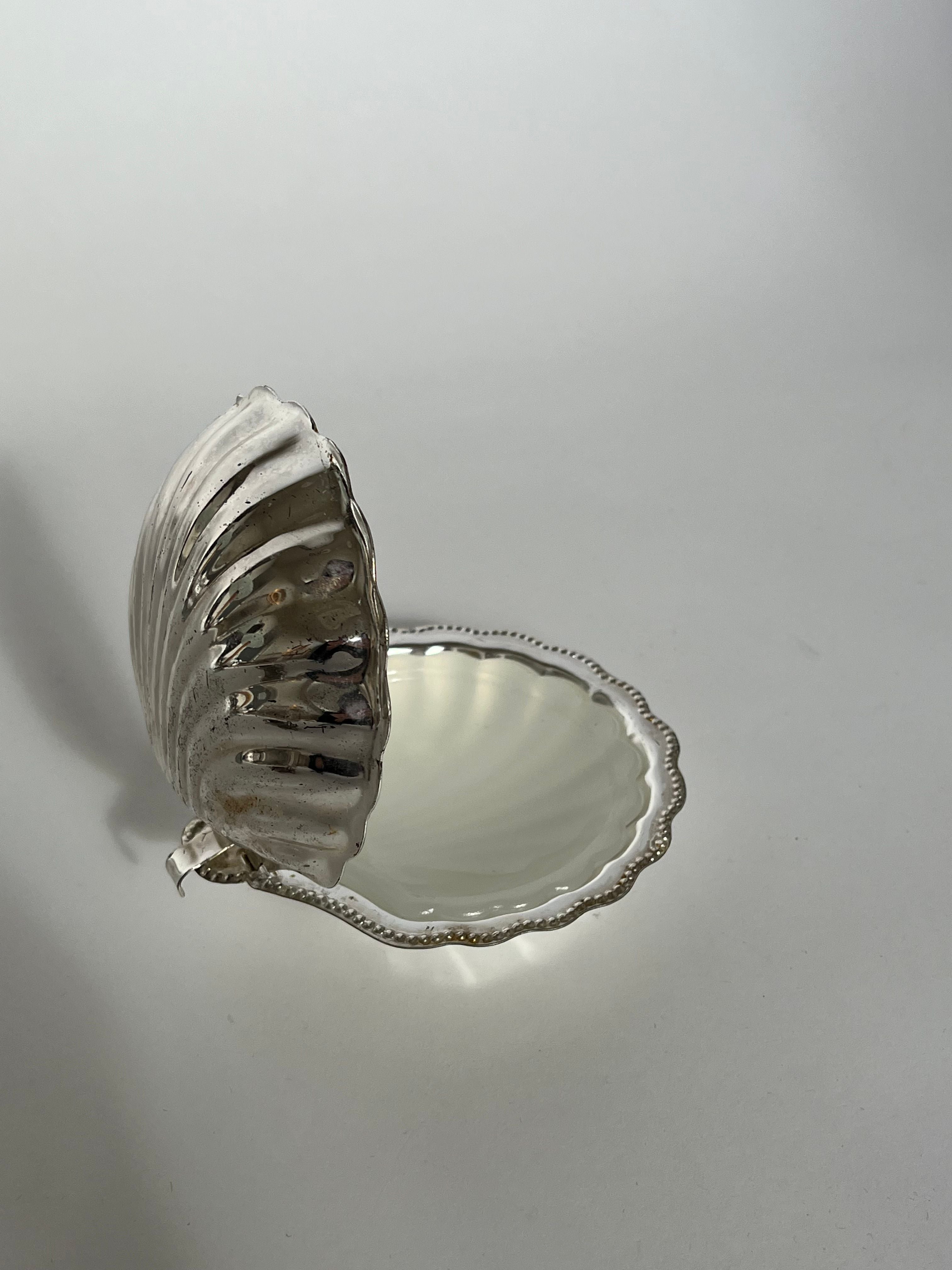 A shell-shaped butter dish from septembre studios, crafted with a vintage brass design and hinged lid slightly ajar, reveals its smooth, reflective interior. The seashell-inspired piece features a scalloped edge and rests elegantly on a gray surface against a plain background.