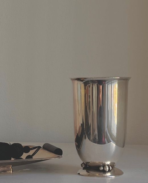 An Art Deco Silver Vase 1940s by Médecine, with a rounded base, sits on a light surface next to a dish of dark grapes and a closed book, set against a plain beige wall.