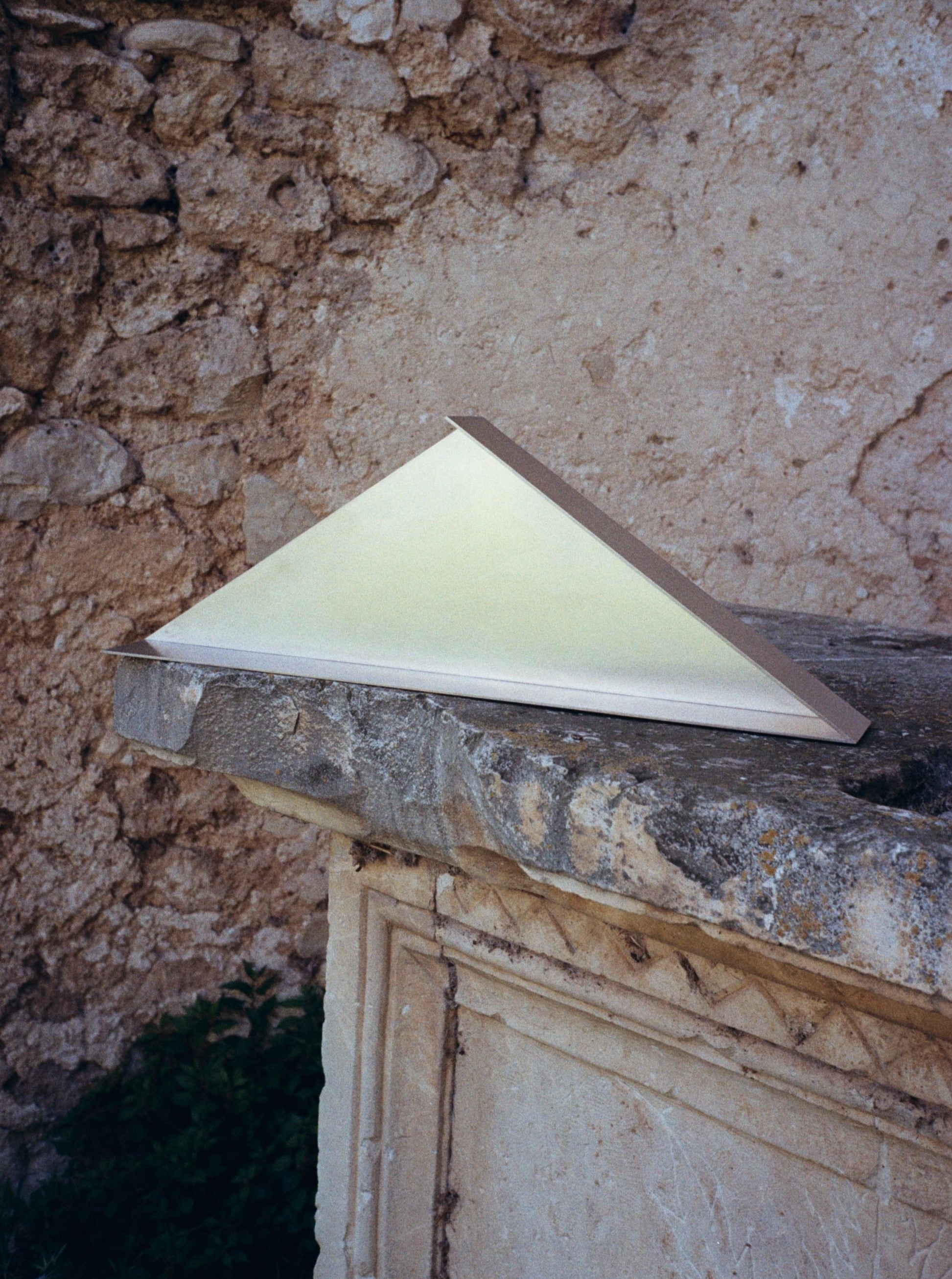 A limited edition triangular prism-shaped light fixture, The Front - Shelf by COSEINCORSO, rests on a ledge against a weathered stone wall. The stainless steel design gives off a warm, soft glow, illuminating part of the stone surface. The setting appears rustic and aged, with natural textures and earth tones.