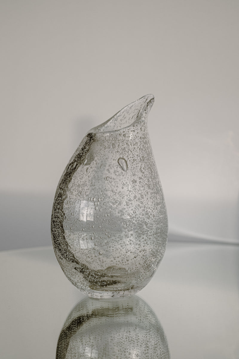 The Small Bubble Vase by septembre studios, featuring a unique, irregular shape and small bubbles enhancing its craftsmanship, sits on a reflective surface against a soft beige background.