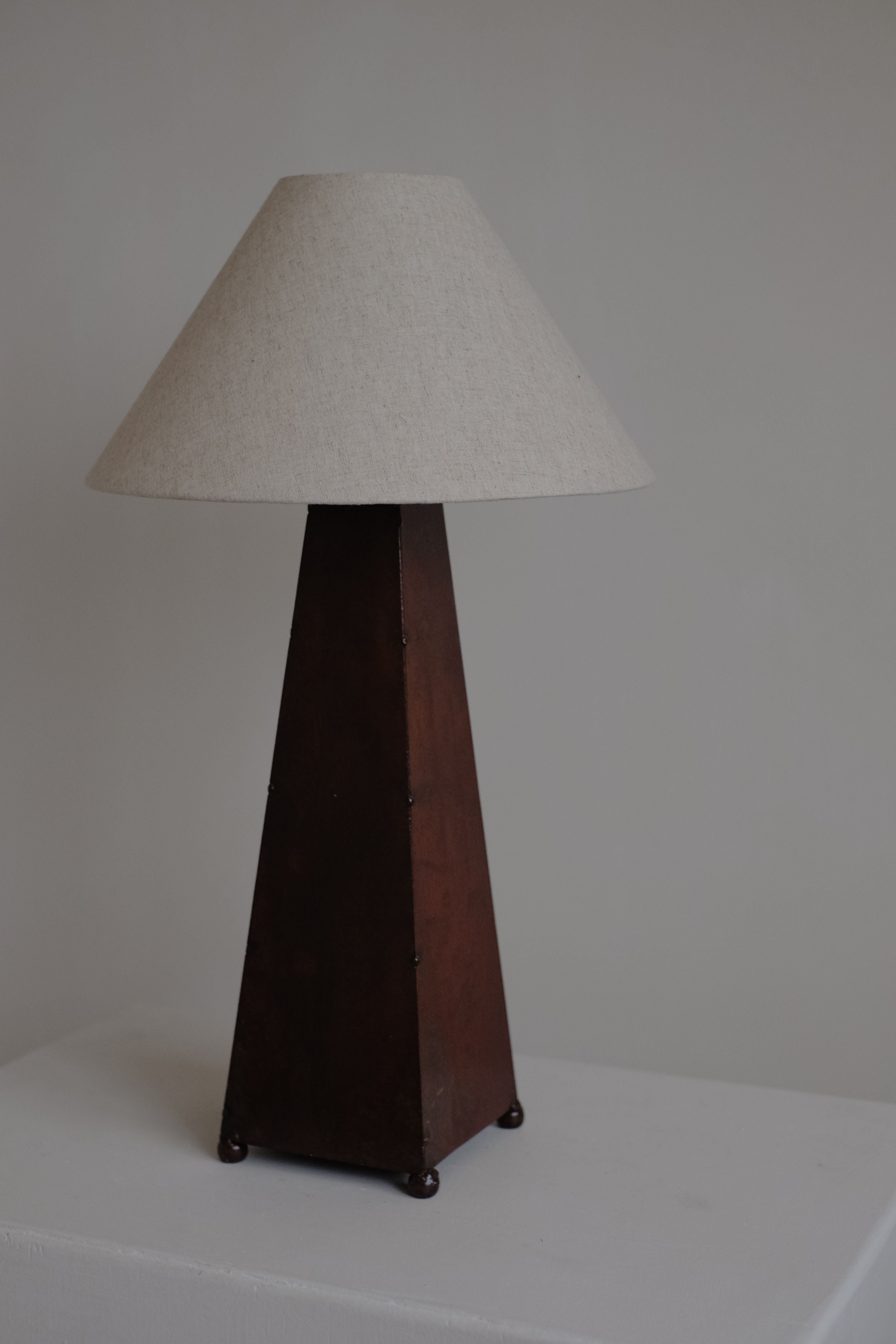 The septembre studios Obelisk Table Lamp has a trapezoidal dark brown wooden base and a beige cone-shaped fabric shade. It sits elegantly on a white surface against a light wall and includes a dimmable switch for ideal lighting.