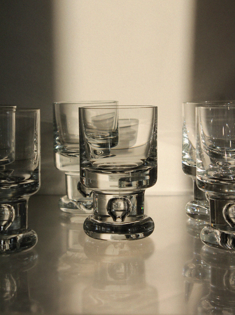 Six empty Tapio Wirkkala Set of Glasses by Vintage Stories are arranged on a reflective surface, with warm sunlight creating dramatic highlights and shadows on the clear mid-century-inspired glass and their reflections.