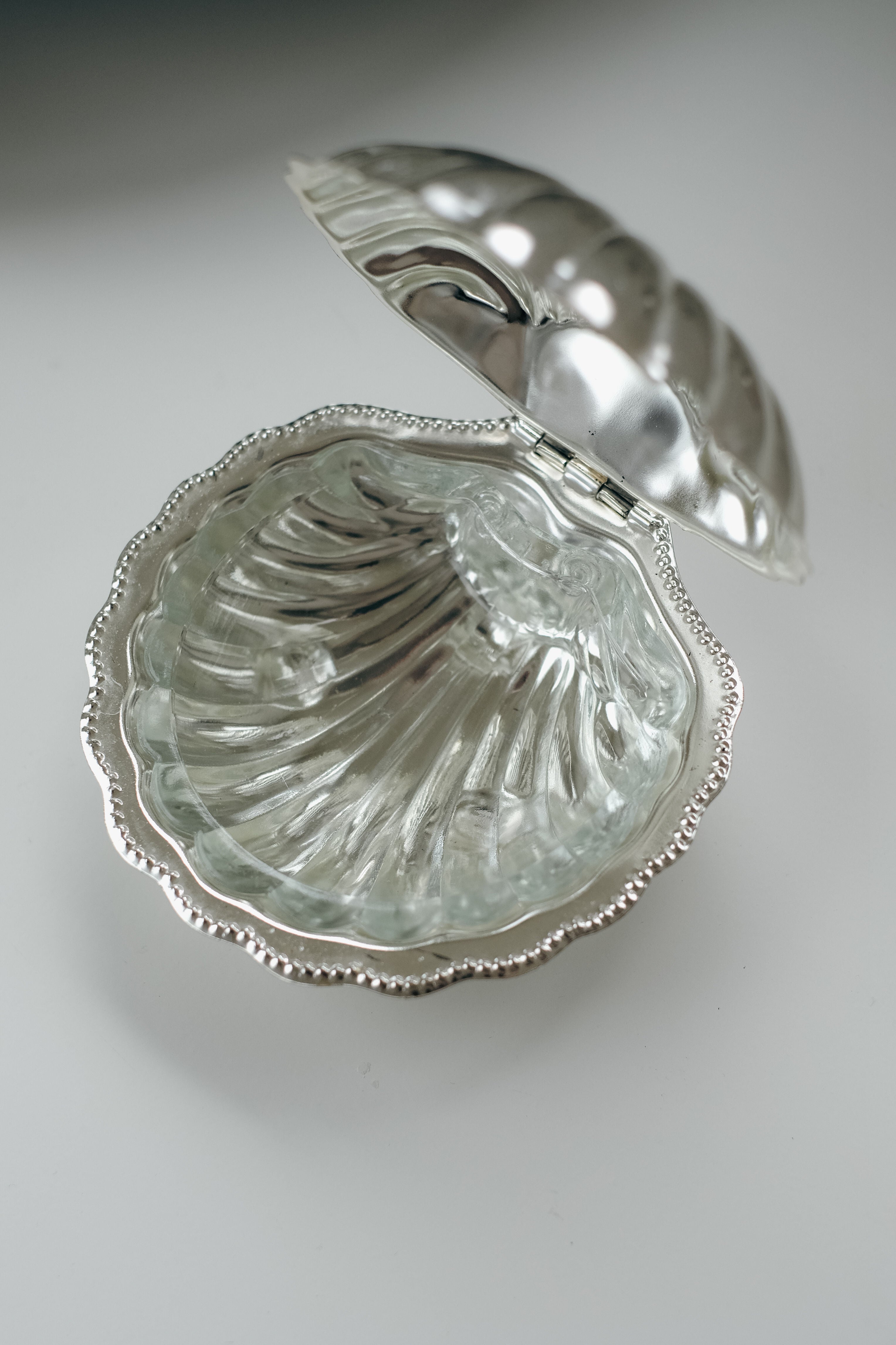 A shiny silver Shell Shaped Butter Dish by septembre studios, resembling a clam with a scalloped edge, is open to display its removable glass insert like a shell's interior, beautifully set against a plain white background.