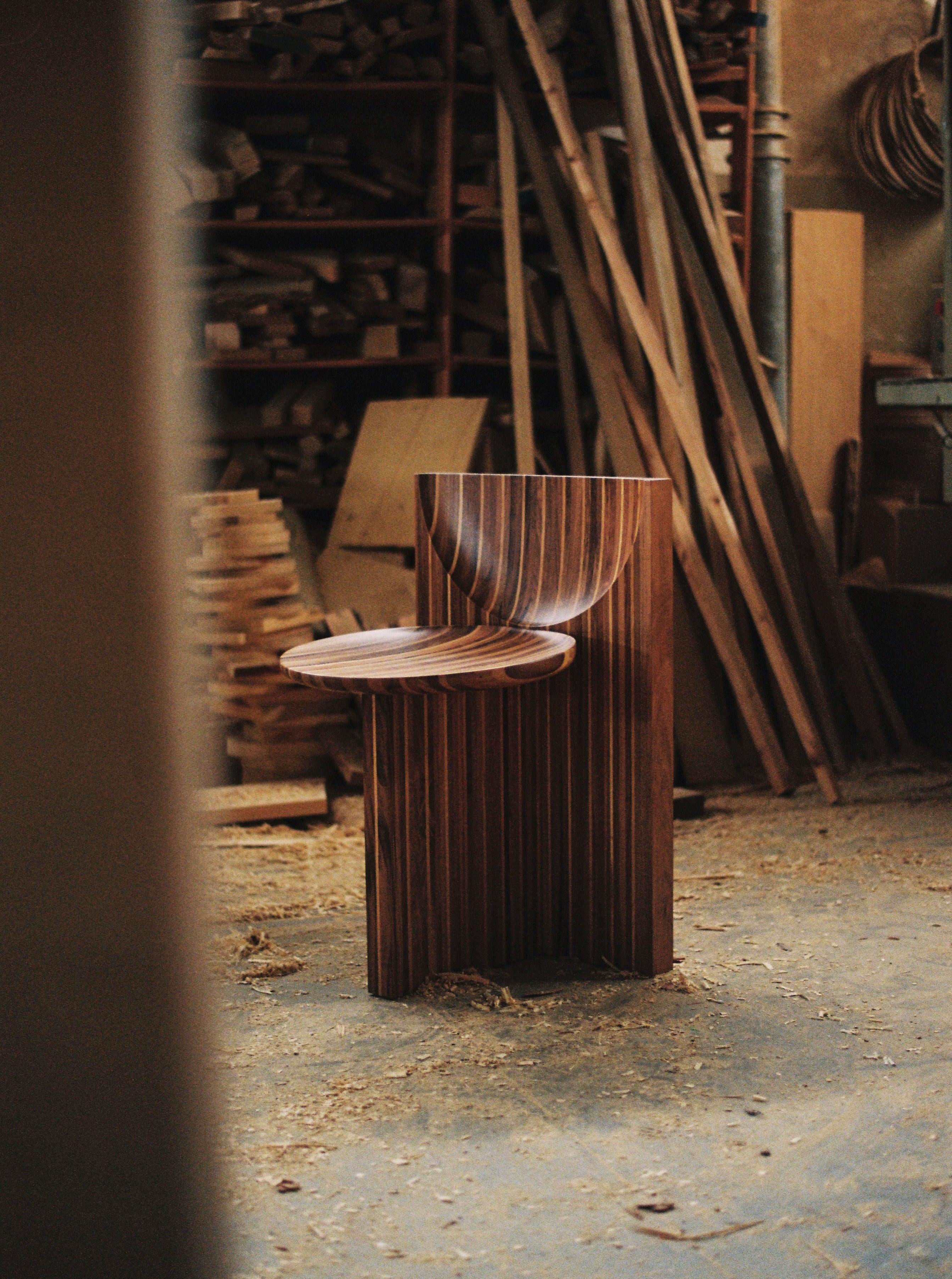 The Vaga Chair by Rosana Sousa, a uniquely designed handcrafted piece made in Portugal, stands in a workshop filled with various wooden planks and woodworking tools. This chair features geometrically intricate patterns, curved lines, and contrasting solid walnut and oak wood tones that highlight its exceptional craftsmanship.