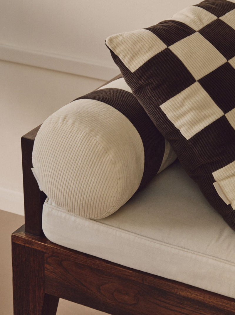 Close-up of a couch with a wooden frame. The armrest features a cylindrical cushion with white and dark brown corduroy stripes, complemented by a black and white checkered throw pillow. Adding character, the sofa includes a vintage-inspired patchwork Handmade UK cushion. The background is softly lit with neutral tones on the wall and floor. Showcased prominently is the AMÉLIORÉ Le Carré - Cotton Corduroy - Chalk/Espresso seat upholstery.