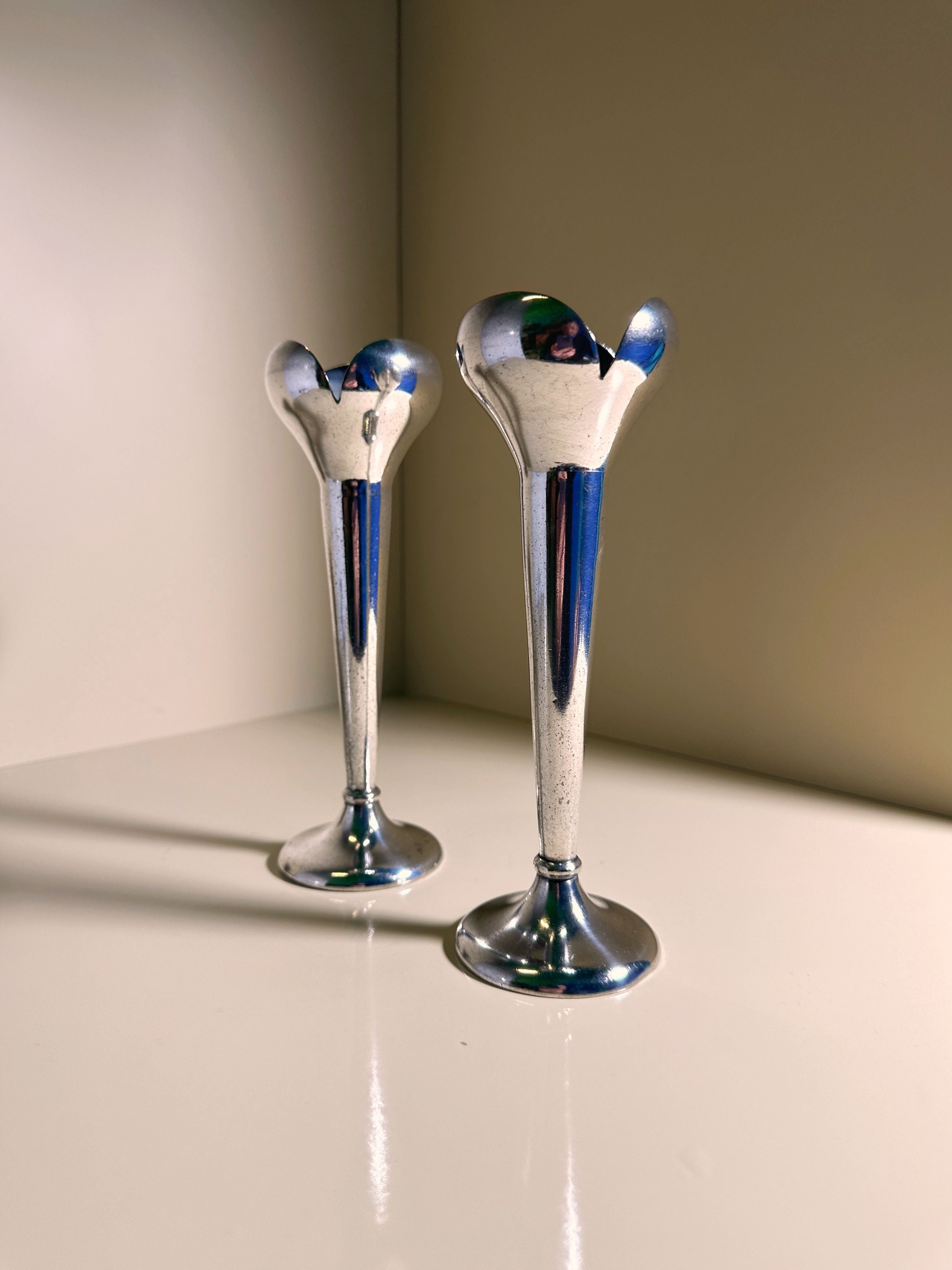 Two reflective Alpacca silver candlesticks by Gio Ponti for Calderoni from the 1930s, reminiscent of trumpet vases with flared tops, sit side by side on a light surface. They cast soft shadows against a muted wall background, embodying graceful elegance. Available at Dodo Vintage.