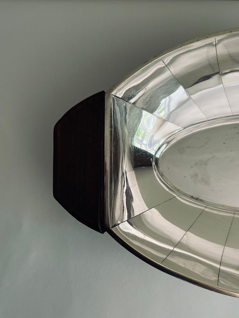 A close-up view of the elegant Art Deco Serving Tray by Flock Studio, featuring an oval shape and dark wooden handles on a gray surface. The polished metal reflects light, highlighting its shiny surface and intricate modernist design.