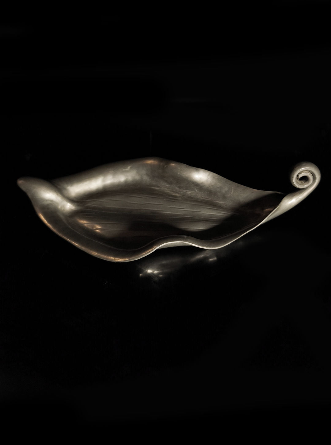 The Handmade Leaf Vide Poche by Les Objoies is a metallic, leaf-shaped decorative tray featuring smooth curves and a spiral tip, set against a black background. Crafted in pewter, it showcases a shiny reflective surface and undulating edges, evoking the essence of an Art Nouveau vide-poche.