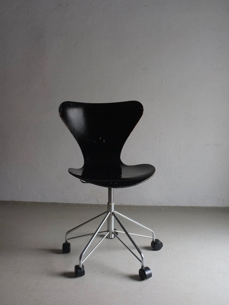 A modern, black office chair with a contoured seat and backrest, reminiscent of an Arne Jacobsen design, is supported by a silver metal base with five caster wheels. Set against a plain gray wall and floor, the sleek and minimalist 3117 Black Swivel Chair by Arne Jacobsen for Fritz Hansen from Veter Vintage updates the classic elegance of a vintage swivel desk chair.