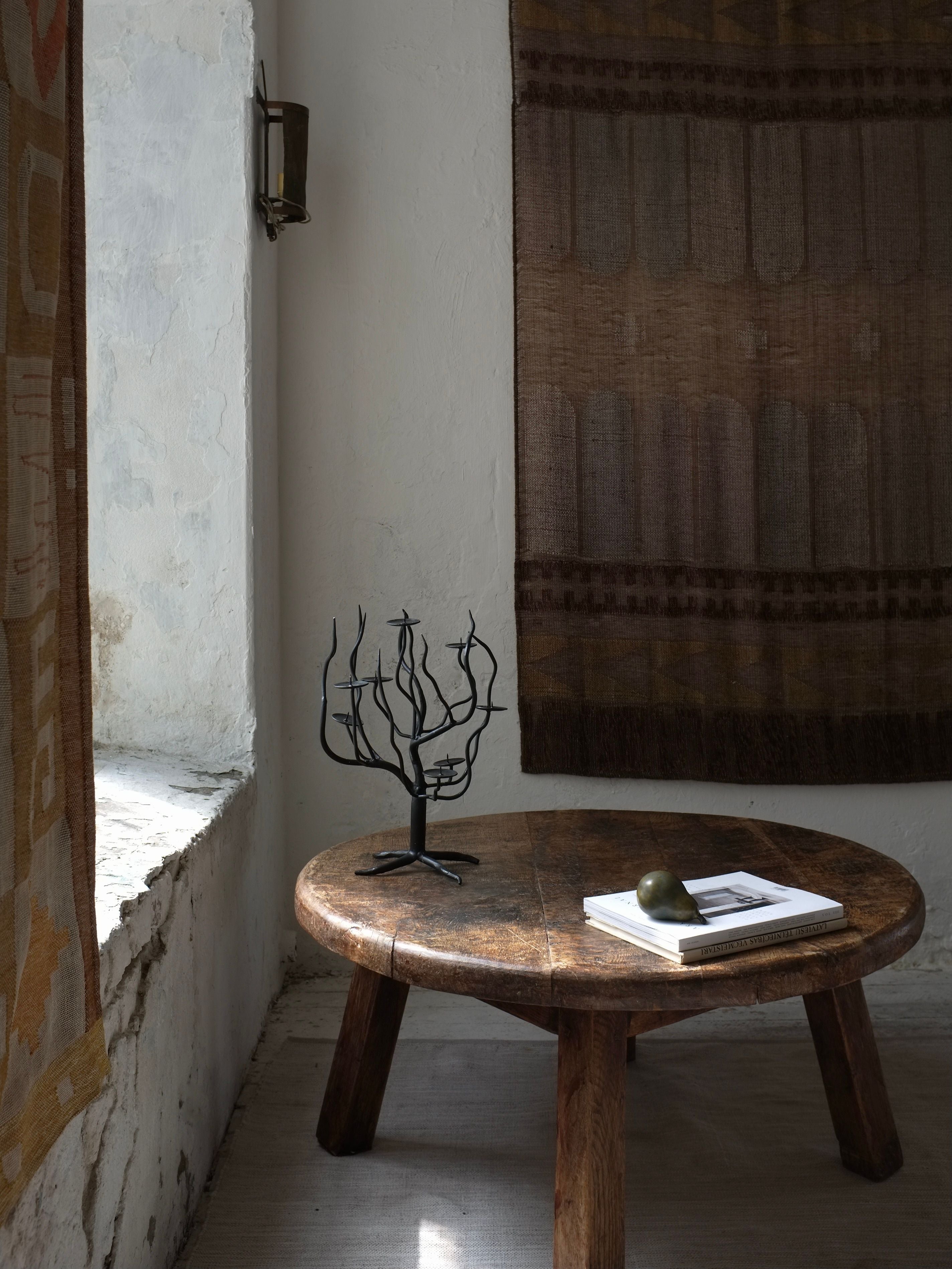 A rustic room highlights a round wooden table adorned with a small, minimalist tree sculpture, a book, and a green pear. The decor is elevated by the elegance of the Hand-forged Metal Sculptural Candle Holder from Veter Vintage. A textured tapestry hangs on the wall while another drapes over part of the window ledge, creating a cozy ambiance. Natural light filters in softly.
