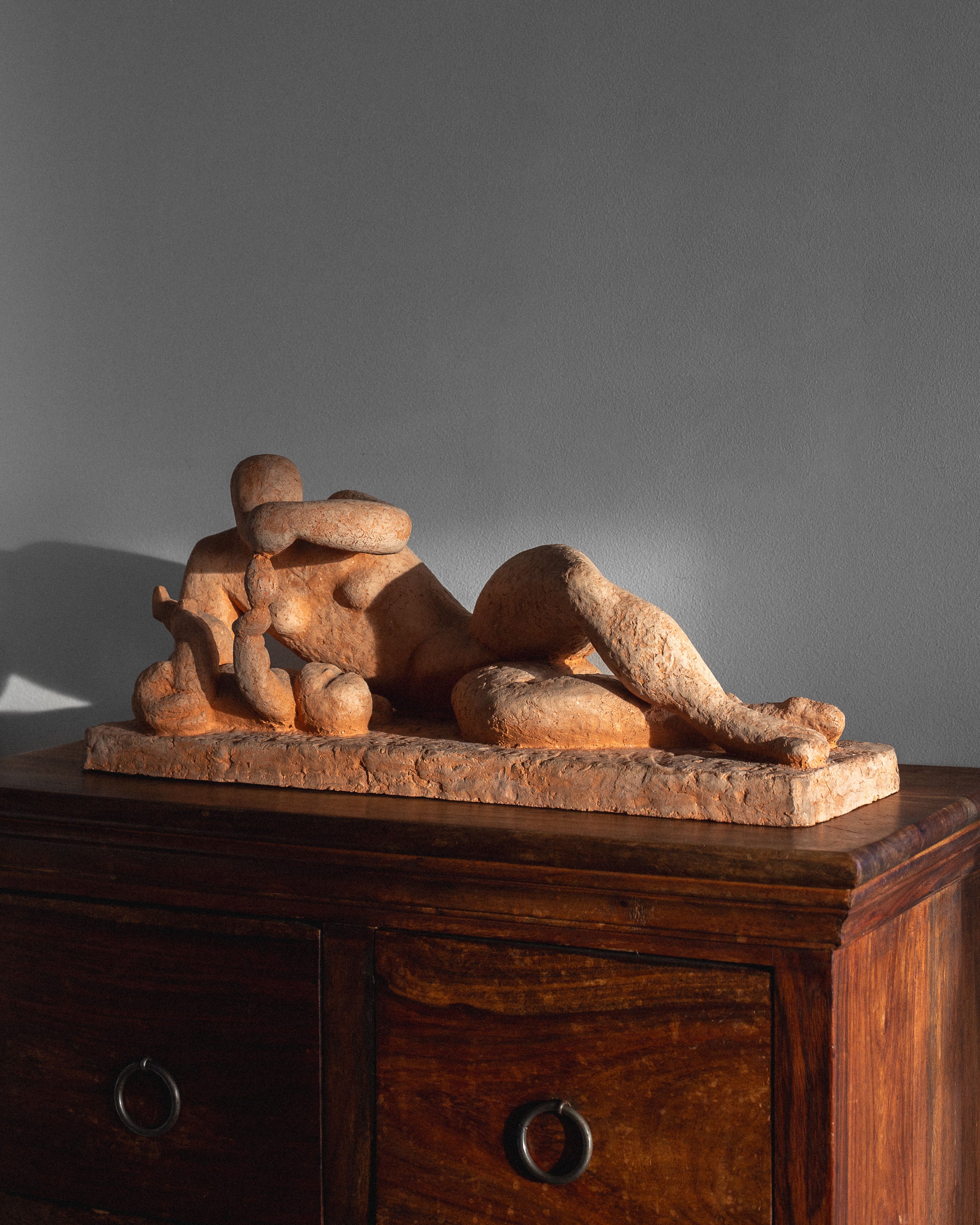 This unglazed terracotta sculpture, "Modernist Terracotta Sculpture by Bengt Amundin, 1950s" from Spigel, showcases a stone figure and child reclining on a wooden furniture piece with drawers. Side lighting creates soft shadows on the grey wall, enhancing its serene elegance.