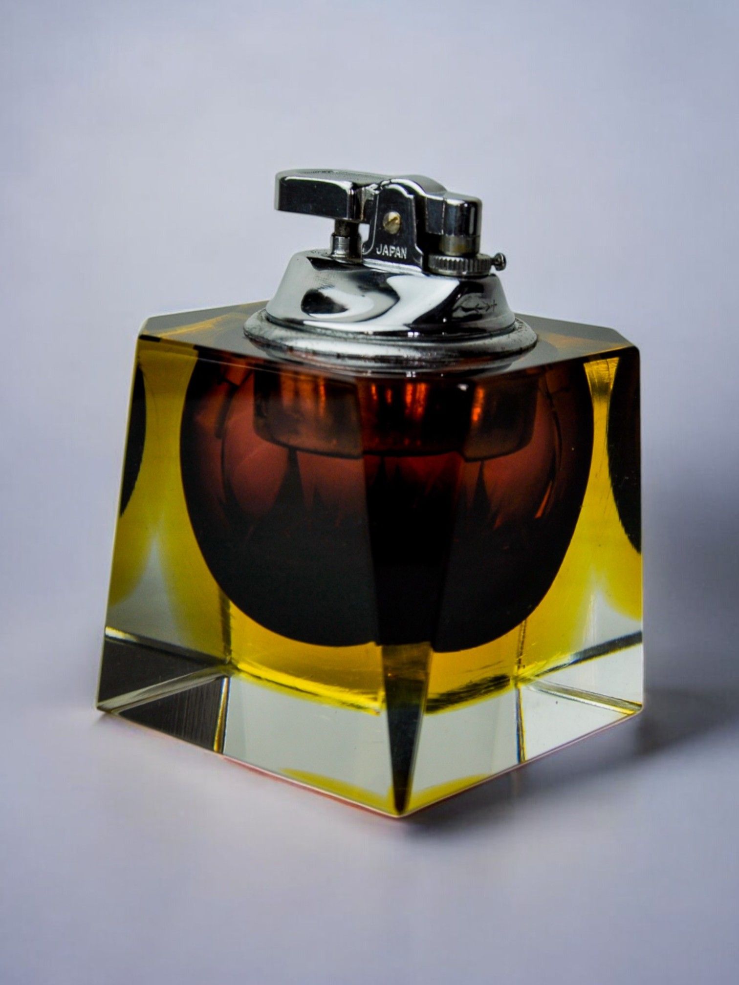 The Sommerso Lighter by Seguso features a brass-toned transparent cubical base reminiscent of sommerso designs, with a metallic top engraved "JAPAN." The Murano glass central container holds dark amber liquid, offering classic elegance. Available exclusively at Malamar Studio.