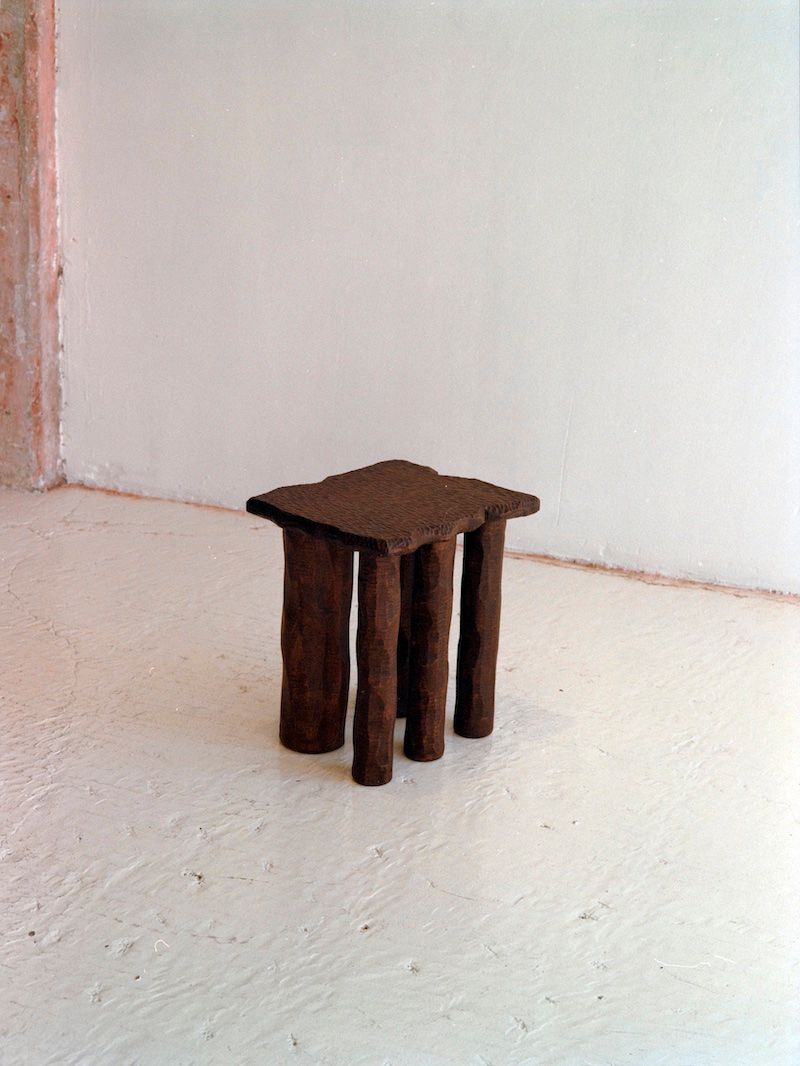 The Mare Side Table by Ombia Studio features four uneven sculpted legs and a rough-textured seat in solid white oak. Positioned on a light floor against an off-white wall, this made-to-order piece has an organic, handcrafted look.