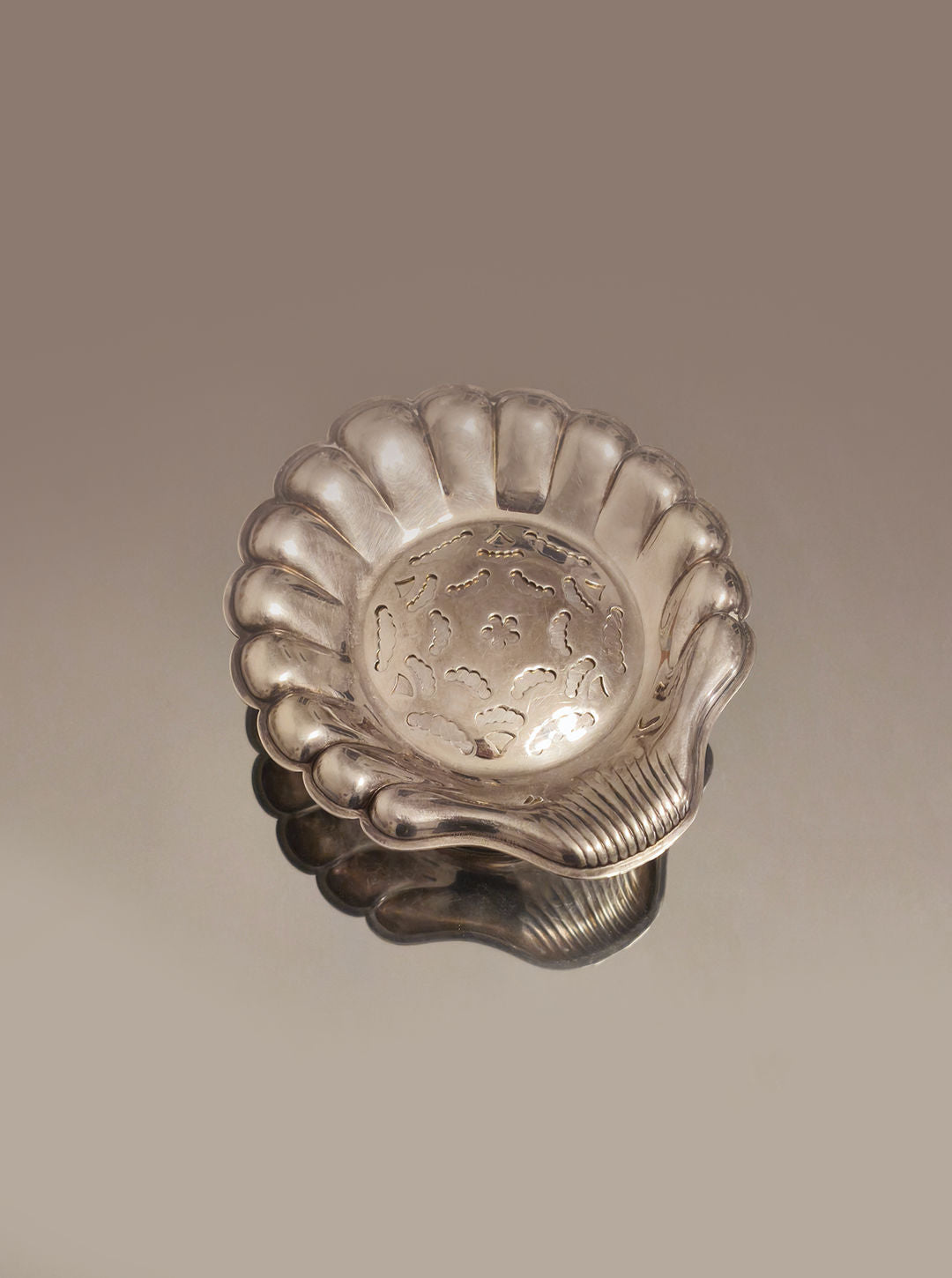 The Shell Butter Dish by Les Objoies is a silver-plated piece with scalloped edges and a seashell-inspired cut-out center, perfect for showcasing butter curls. Its reflective surface adds elegance to any setting.