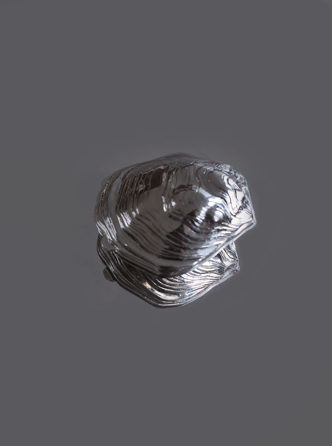 A reflective, metallic object resembling a frog sits on a dark surface, its polished finish showcasing detailed ridges and contours. This intricate piece is the "Small Oyster Box" by Les Objoies, with its textures creating a unique visual effect—an exceptional and unique gift.