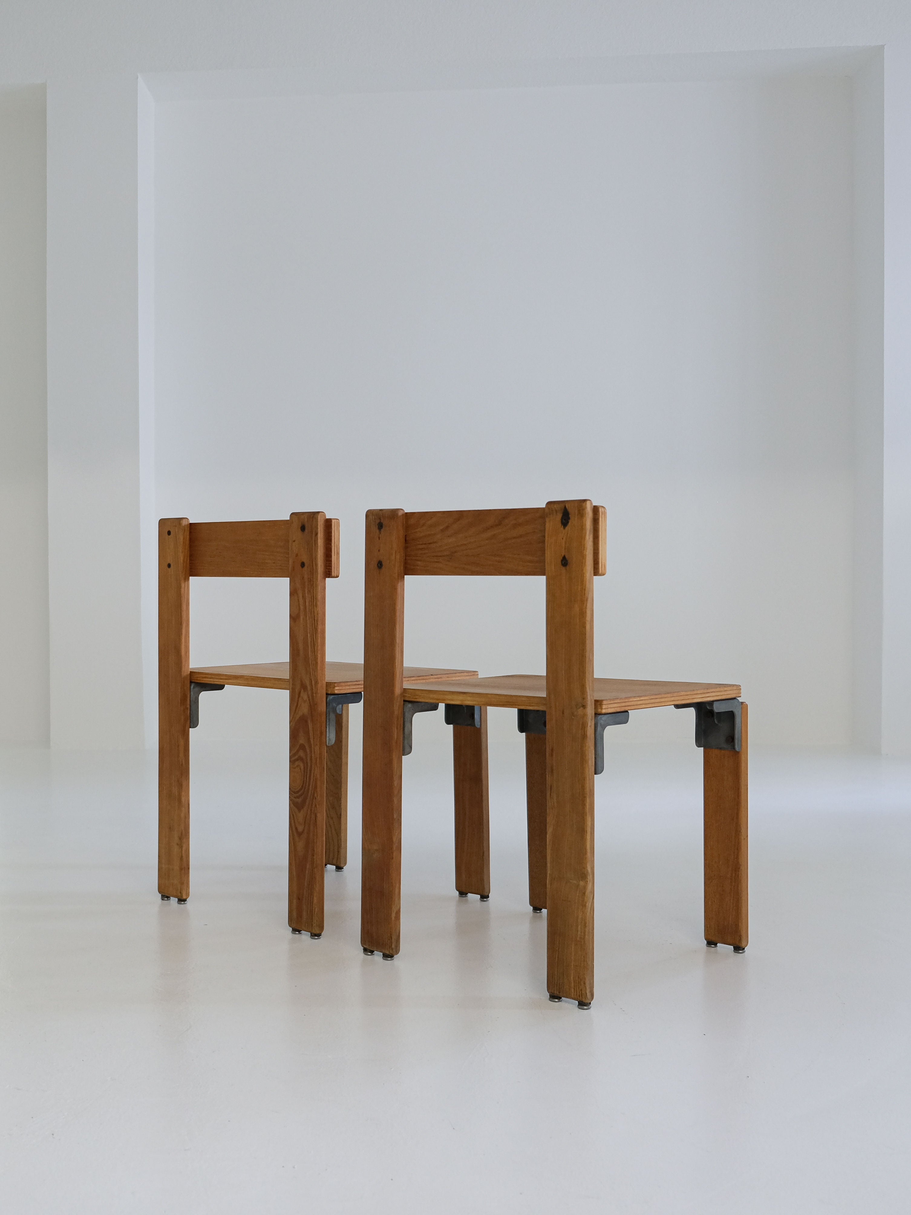 A pair of "Les Carats" dining chairs by George Candilis and Anja Blomstedt from the 1970s, crafted from sleek oak plywood with an angular design, sit on a smooth light floor facing slightly apart against a minimalist white wall. Brand: antibeige.