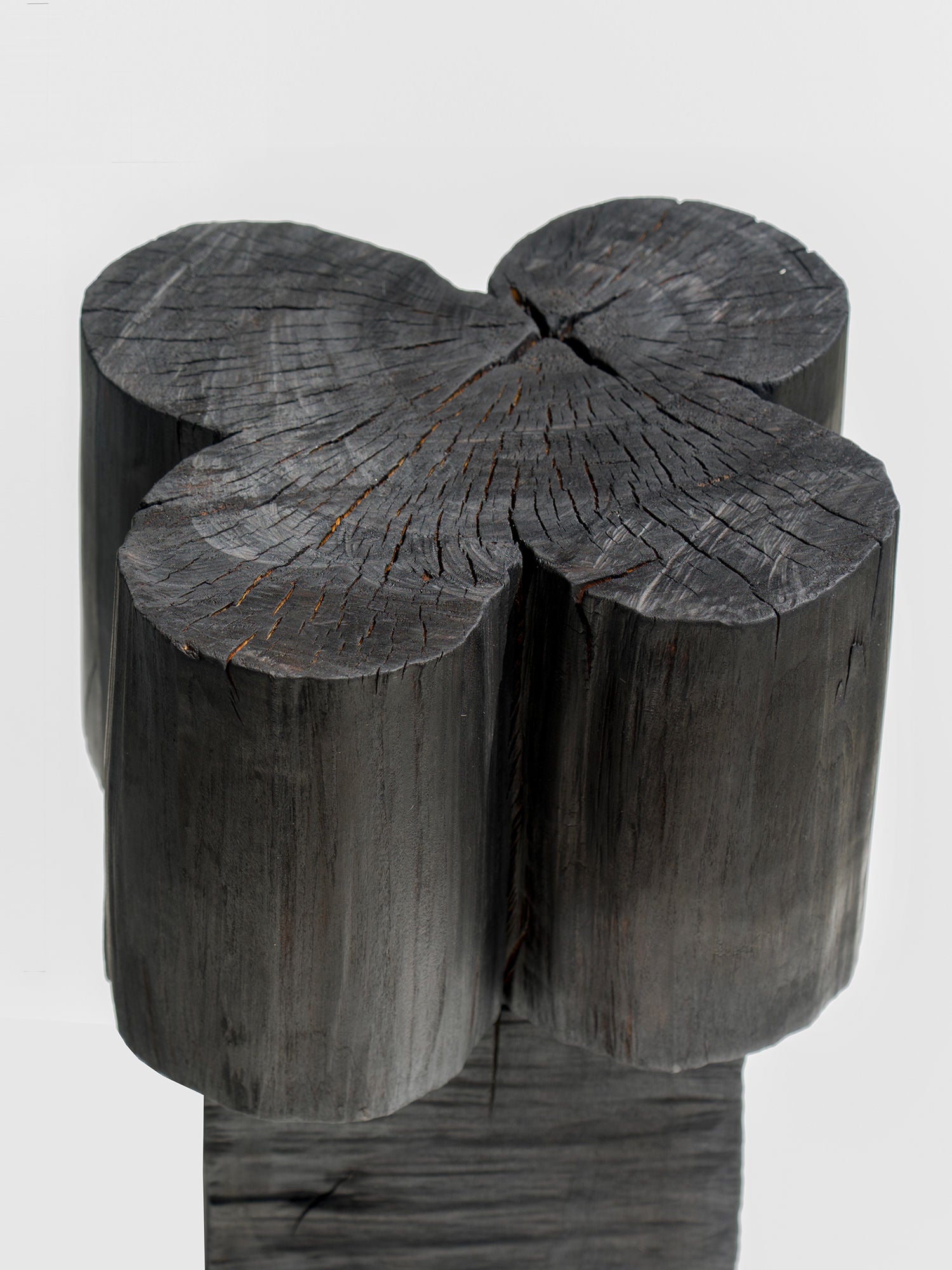 The Burnt Black Coffee Tables/Stools by Touch With Eyes feature a clover-shaped top of four interlocking, charred logs with a cracked and textured surface that beautifully highlights the natural wood grain and darkened finish.