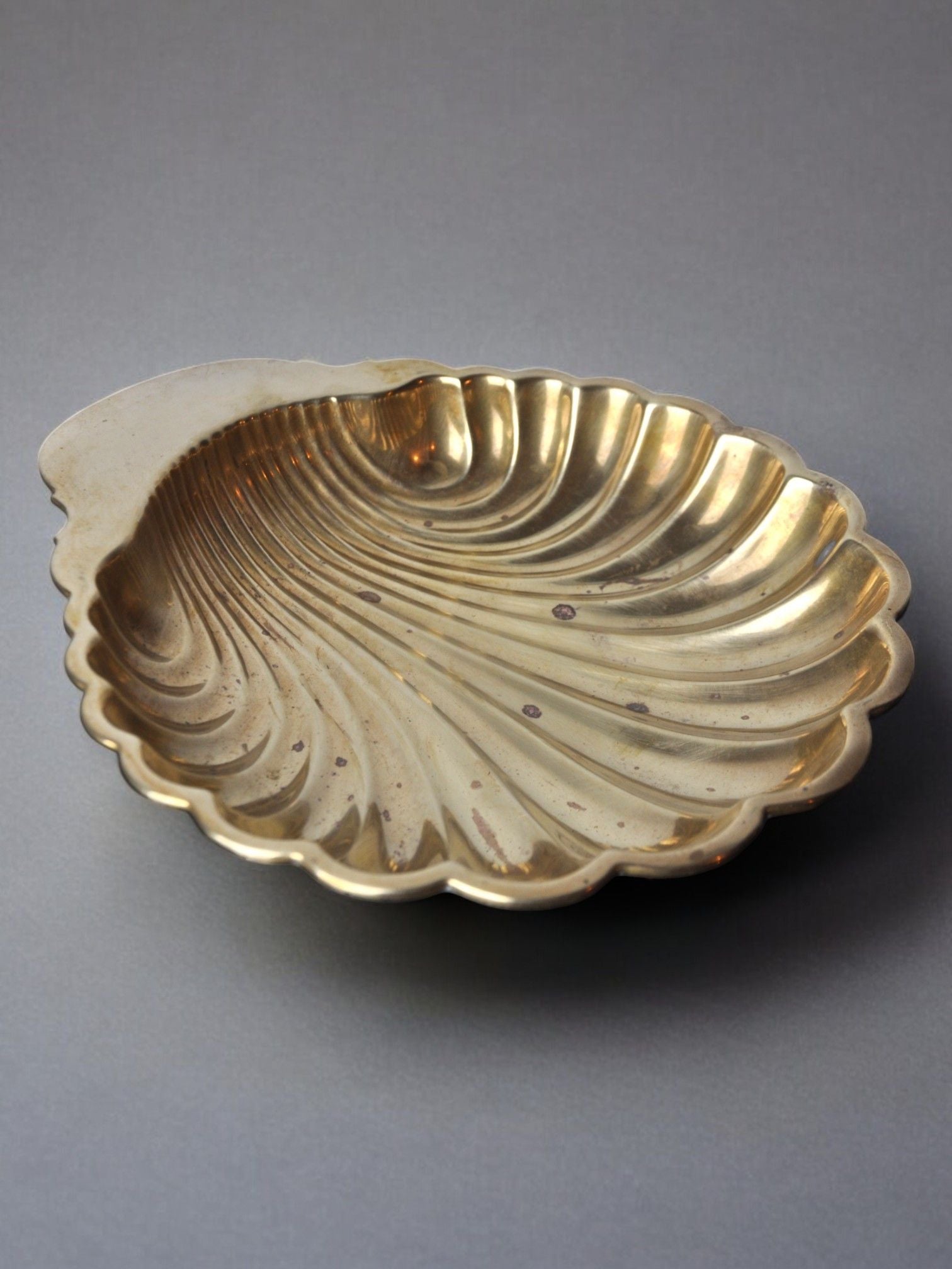 The "Large Shell Shaped Vide-Poche 70s" by Malamar Studio, crafted in Italy from brass and designed to resemble a scallop shell with intricate ridged details, rests on a gray surface. Its gleaming, reflective finish is adorned with a few small tarnish spots, adding to its vintage charm.