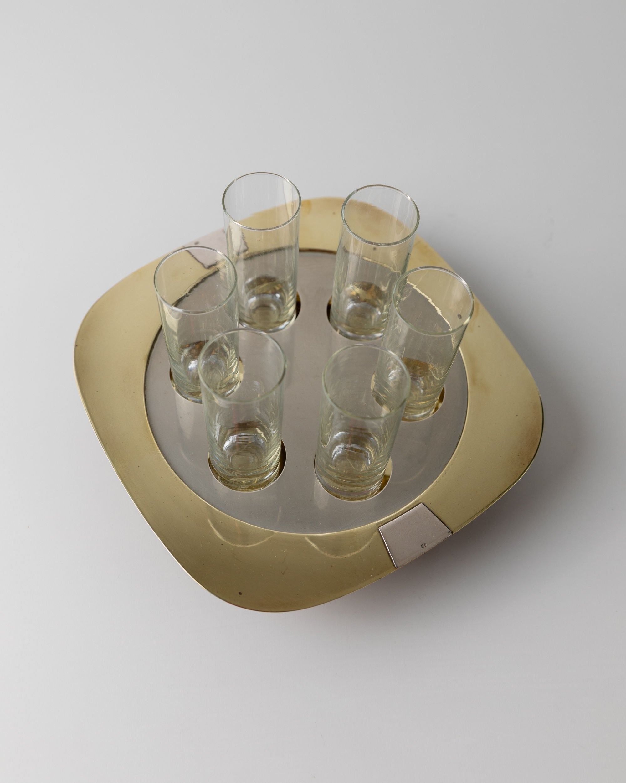 The Bottega Jacobs Set Shot Glasses and Tray showcases a mid-century 70s design, with six clear vintage shot glasses arranged circularly on a square silver-gold tray. The display stands elegantly against a light grey background, enhancing its decorative appeal.