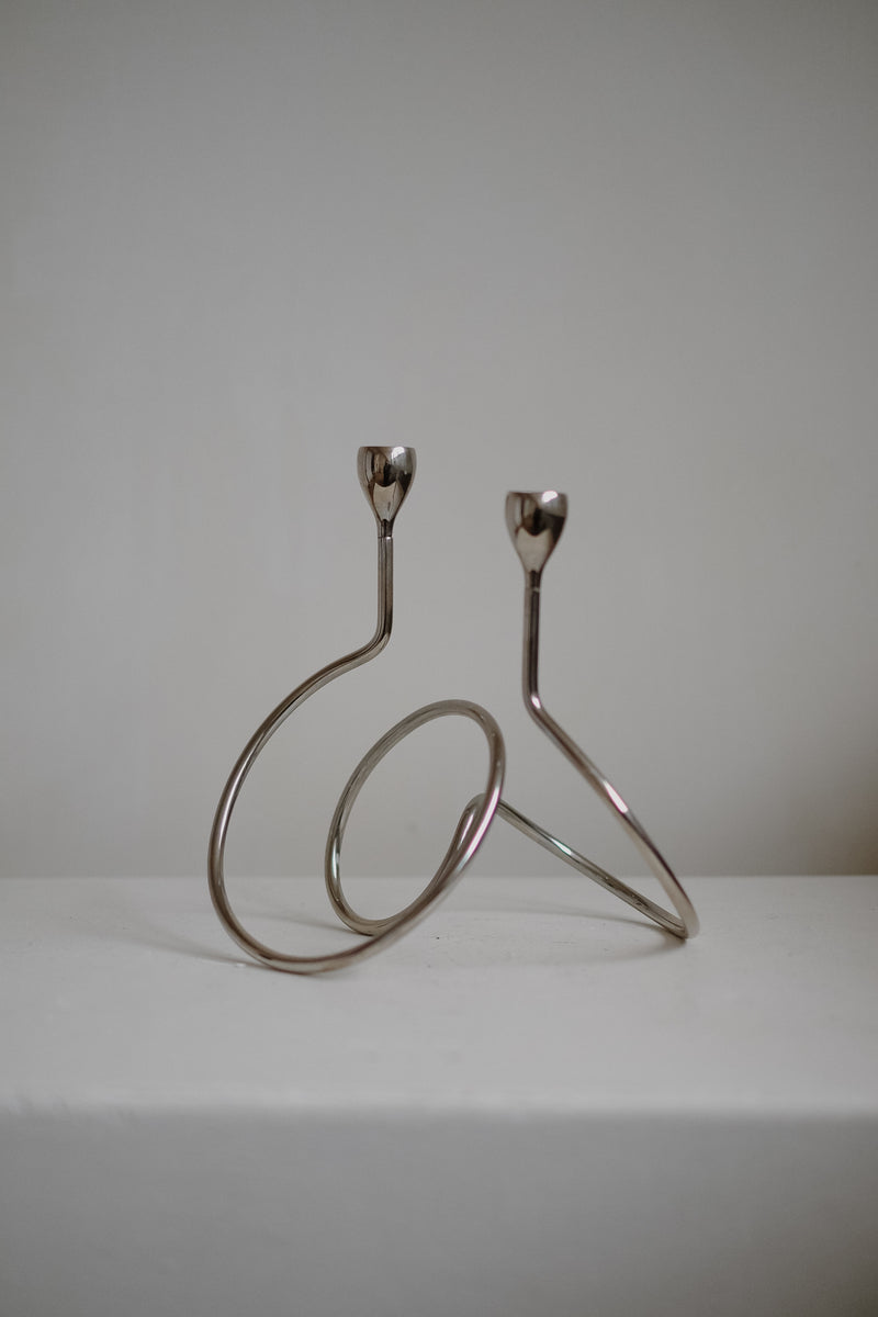 The "Loop" Candle Holder by septembre studios features a minimalist chrome design with two holders at varying heights, ideal for dinner candles. It stands elegantly on a white surface with a simple background, showcasing its artistic, abstract shape.