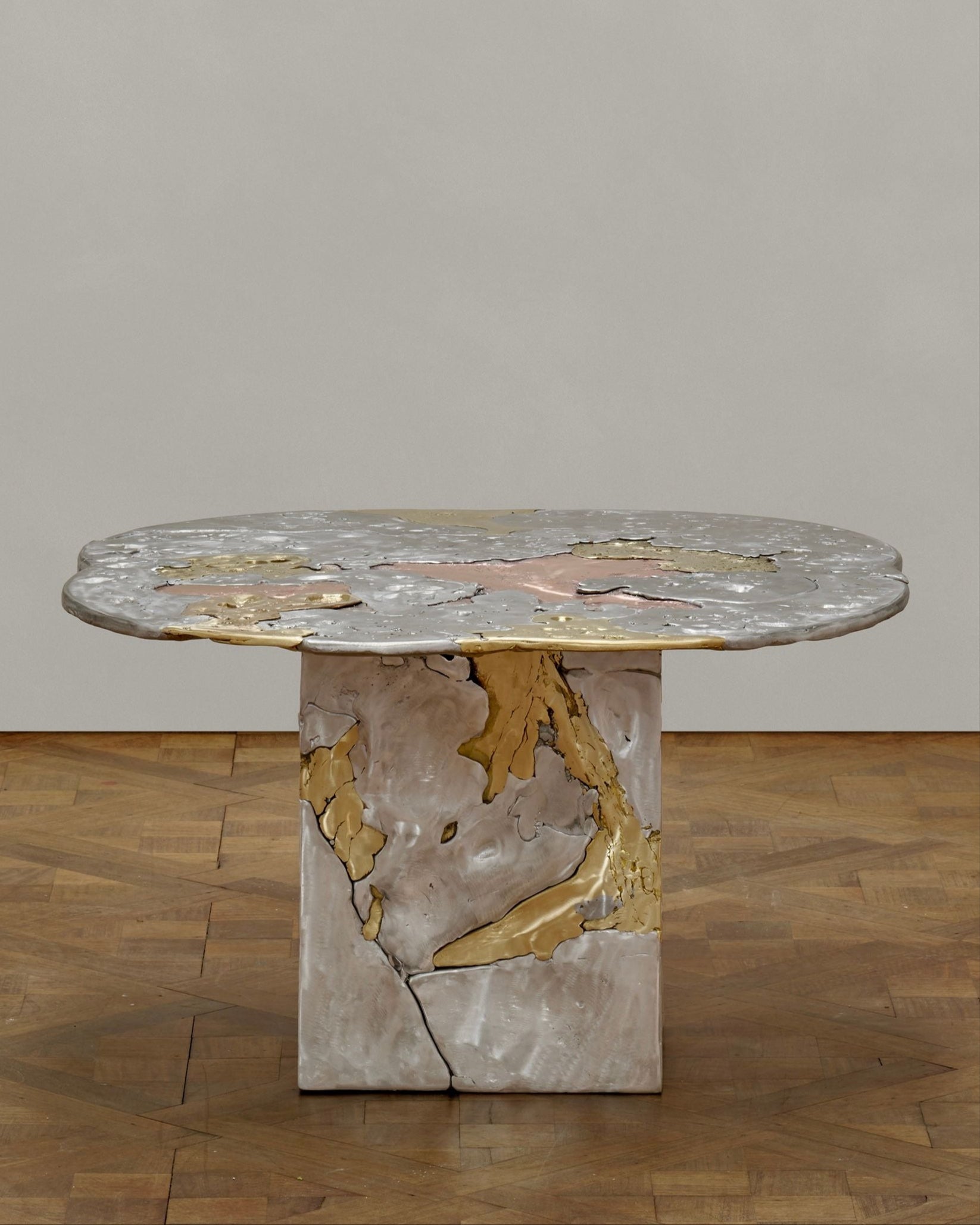 The ‘Elemental Patchwork’ Coffee Table by Sheyang Li features a round marble top with gold and gray veining and a thick pedestal base, shown on parquet flooring against a plain light gray wall.